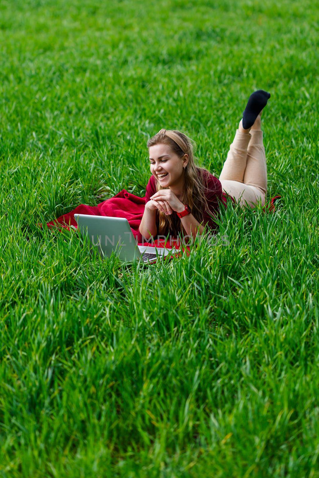 Beautiful young blonde woman is lying on the green grass in the park with a laptop and working. Blue sky with clouds. The girl smiles and enjoys a good day. Work on the nature on a sunny day.