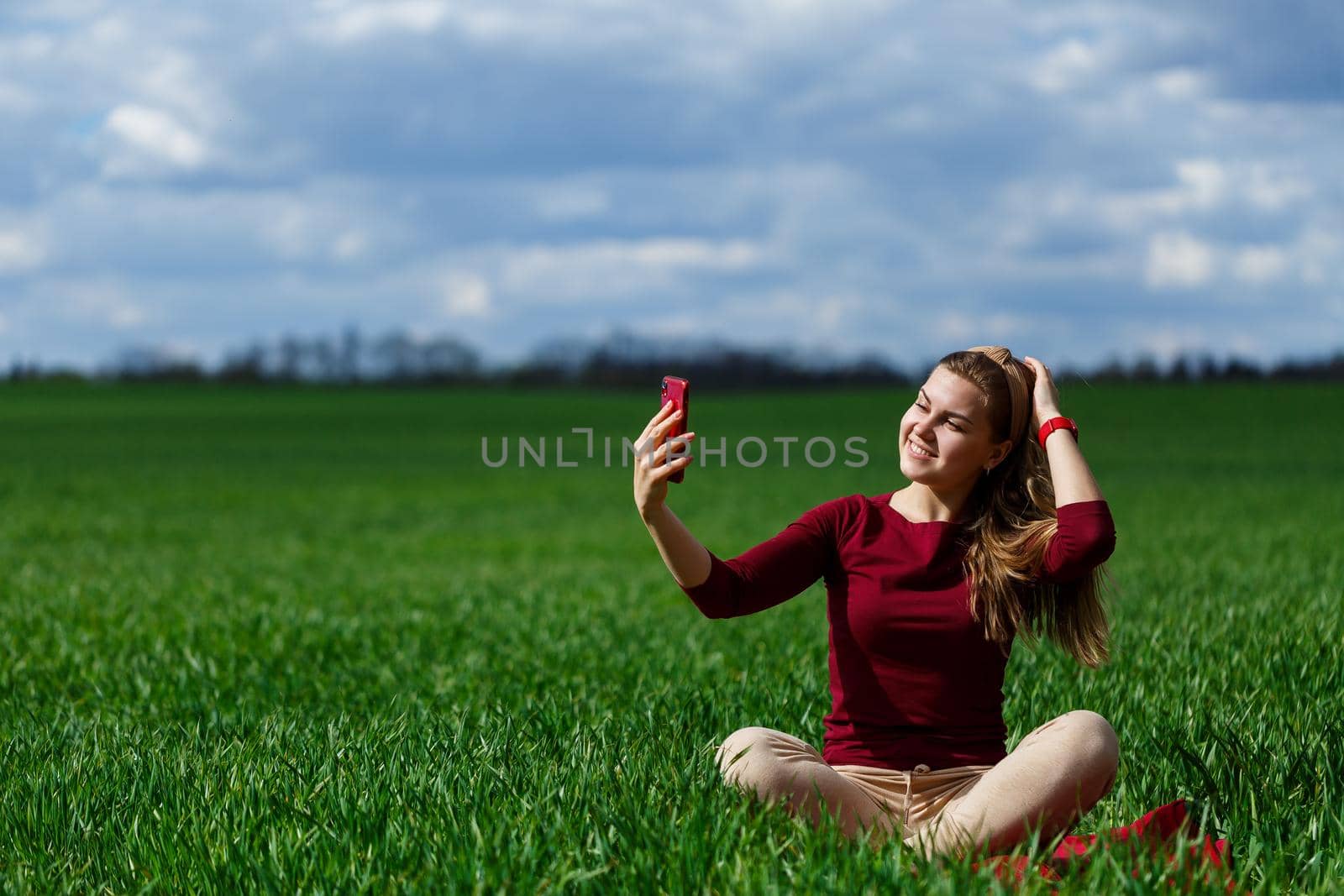 Young beautiful woman student with a phone in her hands sitting on the grass. Girl takes selfies and takes selfie pictures. She smiles and enjoys a warm day. Concept photo on smartphone