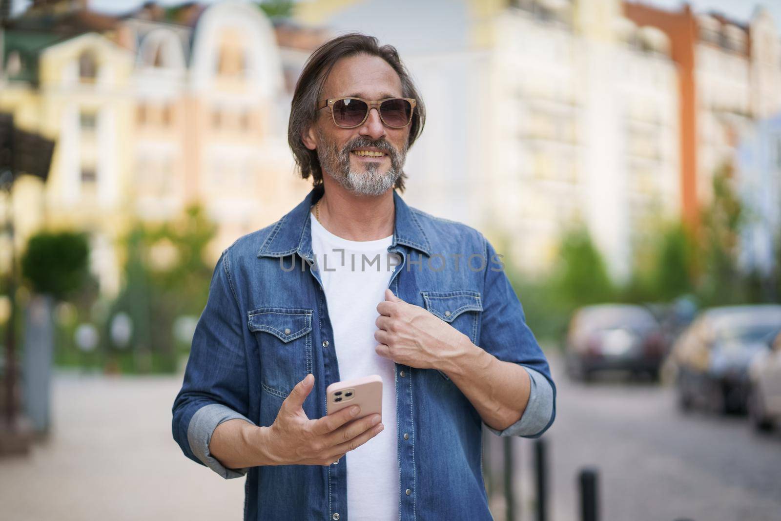 Mature handsome man with smartphone in hand walking on old town streets wearing denim shirt and sunglasses. Handsome middle aged man with grey hair making photos while traveling.