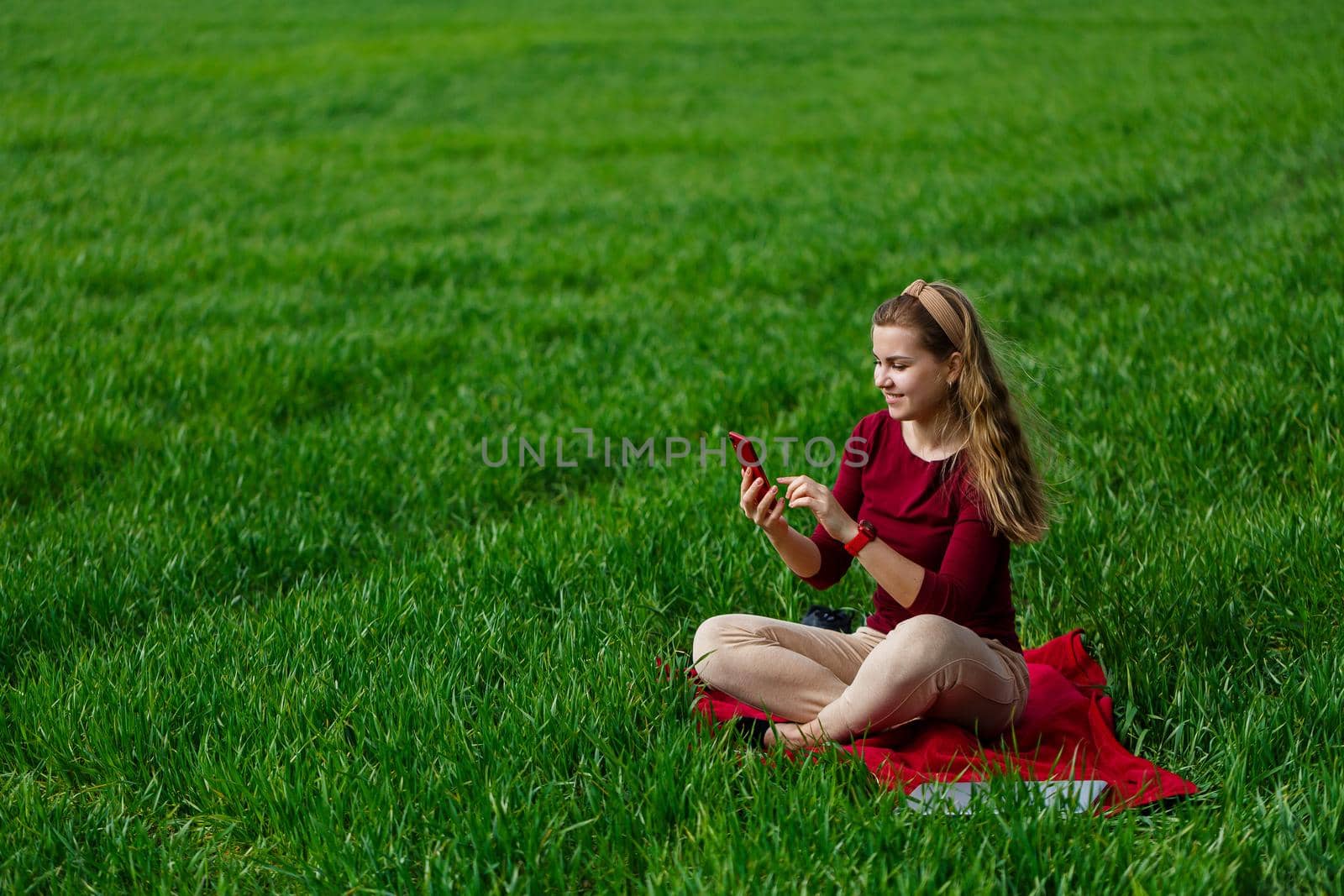 Beautiful woman with a phone in her hands sits on the grass. The girl looks at herself in the camera of the phone and takes selfie photos. She smiles and enjoys a warm day. Concept photo on smartphone
