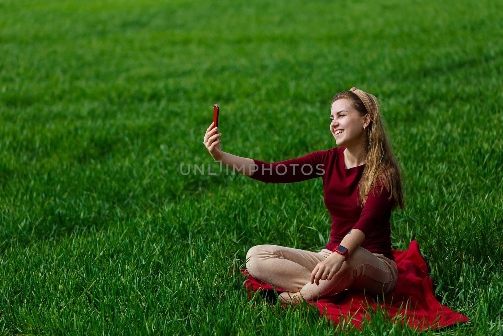 Beautiful woman with a phone in her hands sits on the grass. The girl looks at herself in the camera of the phone and takes selfie photos. She smiles and enjoys a warm day. Concept photo on smartphone