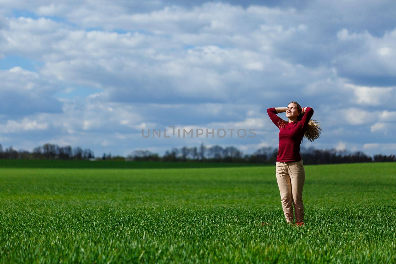 Beautiful young blonde woman stands on green grass in a park. Blue sky with clouds. The girl smiles and enjoys a good day.