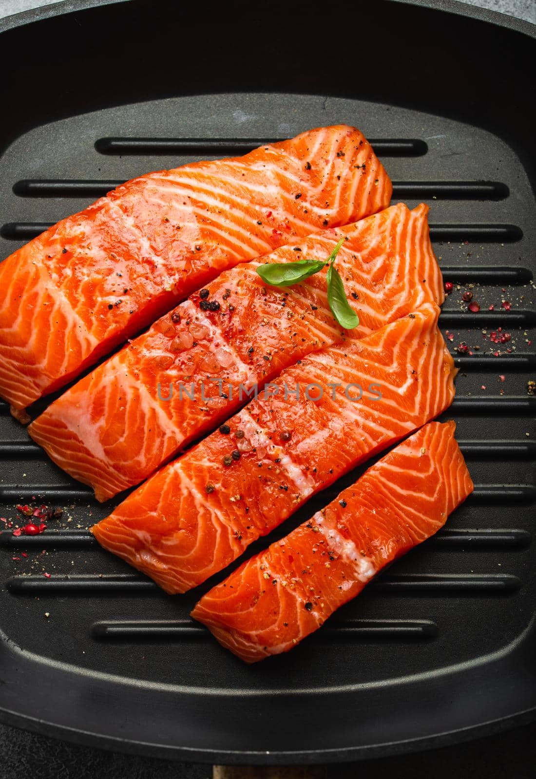 Top view, close-up of cut in slices fresh raw salmon fillet garnished with green basil leaves on grill skillet, gray stone background. Preparing salmon fillet for cooking, healthy eating concept