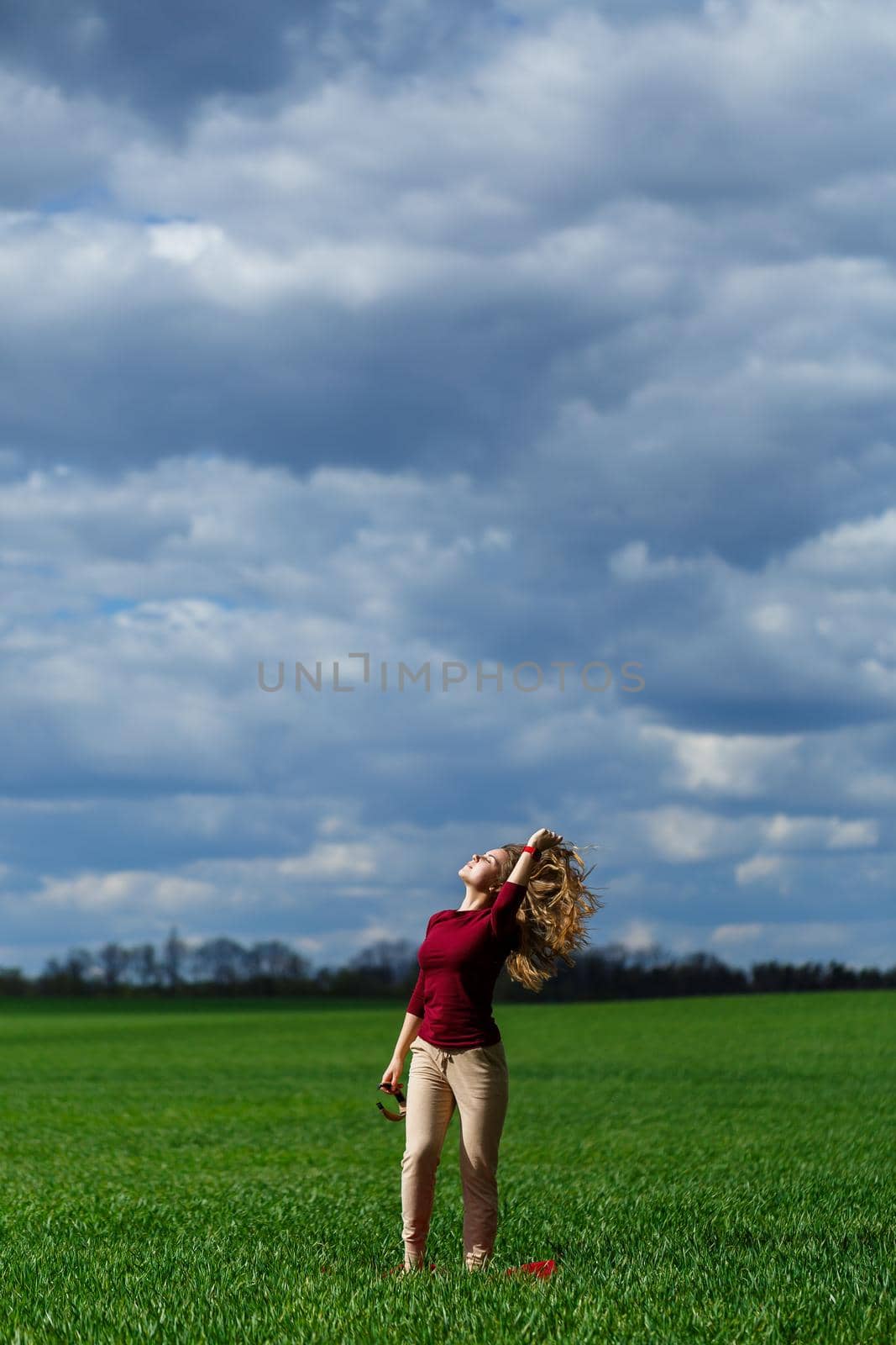 Beautiful young blonde woman stands on green grass in a park. Blue sky with clouds. The girl smiles and enjoys a good day.