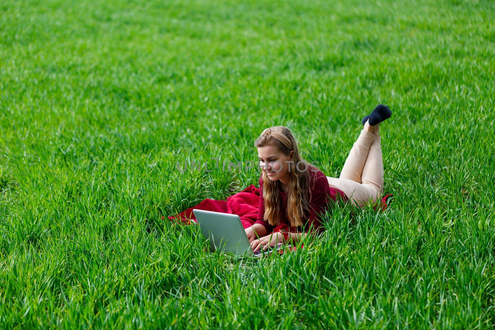 Beautiful young blonde woman is lying on the green grass in the park with a laptop and working. Blue sky with clouds. The girl smiles and enjoys a good day. Work on the nature on a sunny day. by Dmitrytph