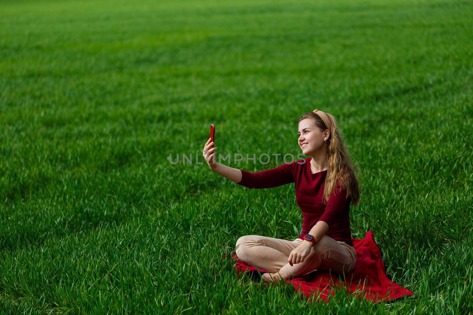 Beautiful woman with a phone in her hands sits on the grass. The girl looks at herself in the camera of the phone and takes selfie photos. She smiles and enjoys a warm day. Concept photo on smartphone
