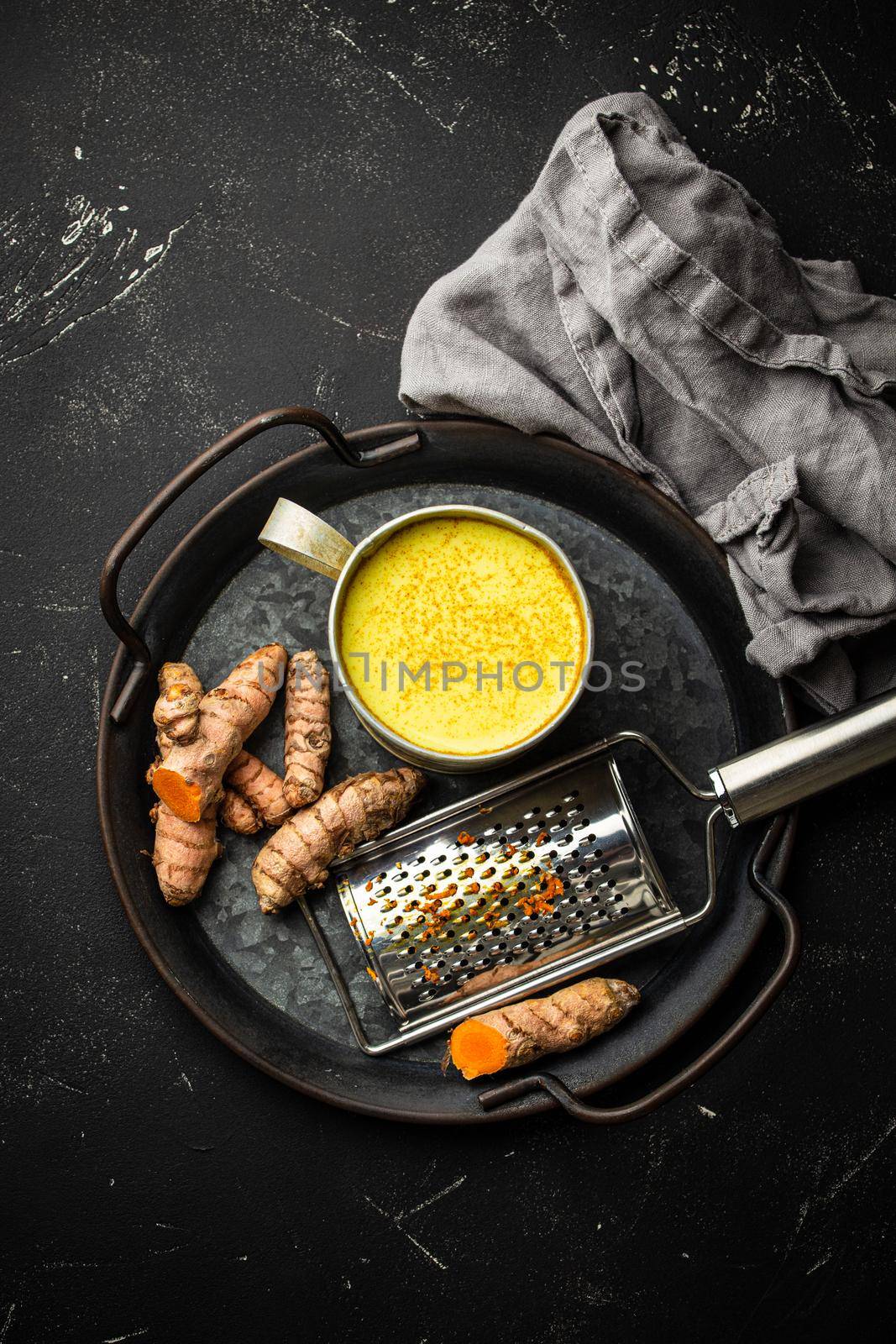 Healthy warm beverage for boosting immune system, turmeric golden milk. Ingredients for cooking detox curcuma milk in rustic tin cup, fresh turmeric root, grater on black stone background, top view.