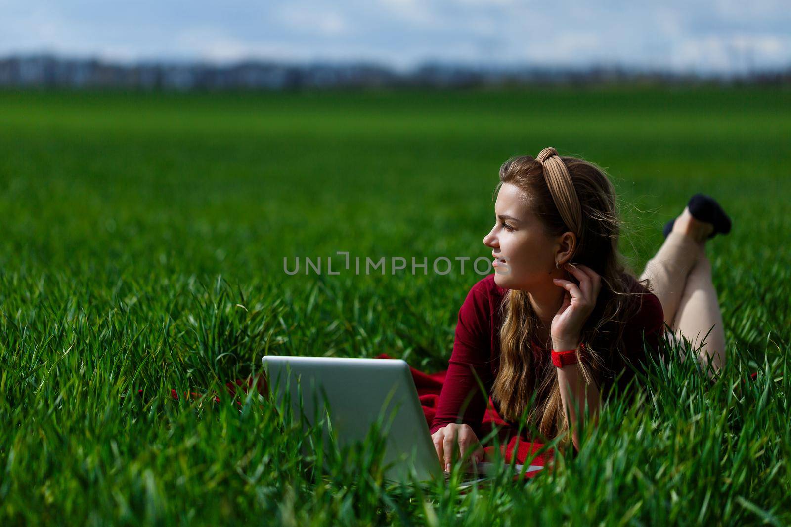 Beautiful young blonde woman is lying on the green grass in the park with a laptop and working. Blue sky with clouds. The girl smiles and enjoys a good day. Work on the nature on a sunny day. by Dmitrytph