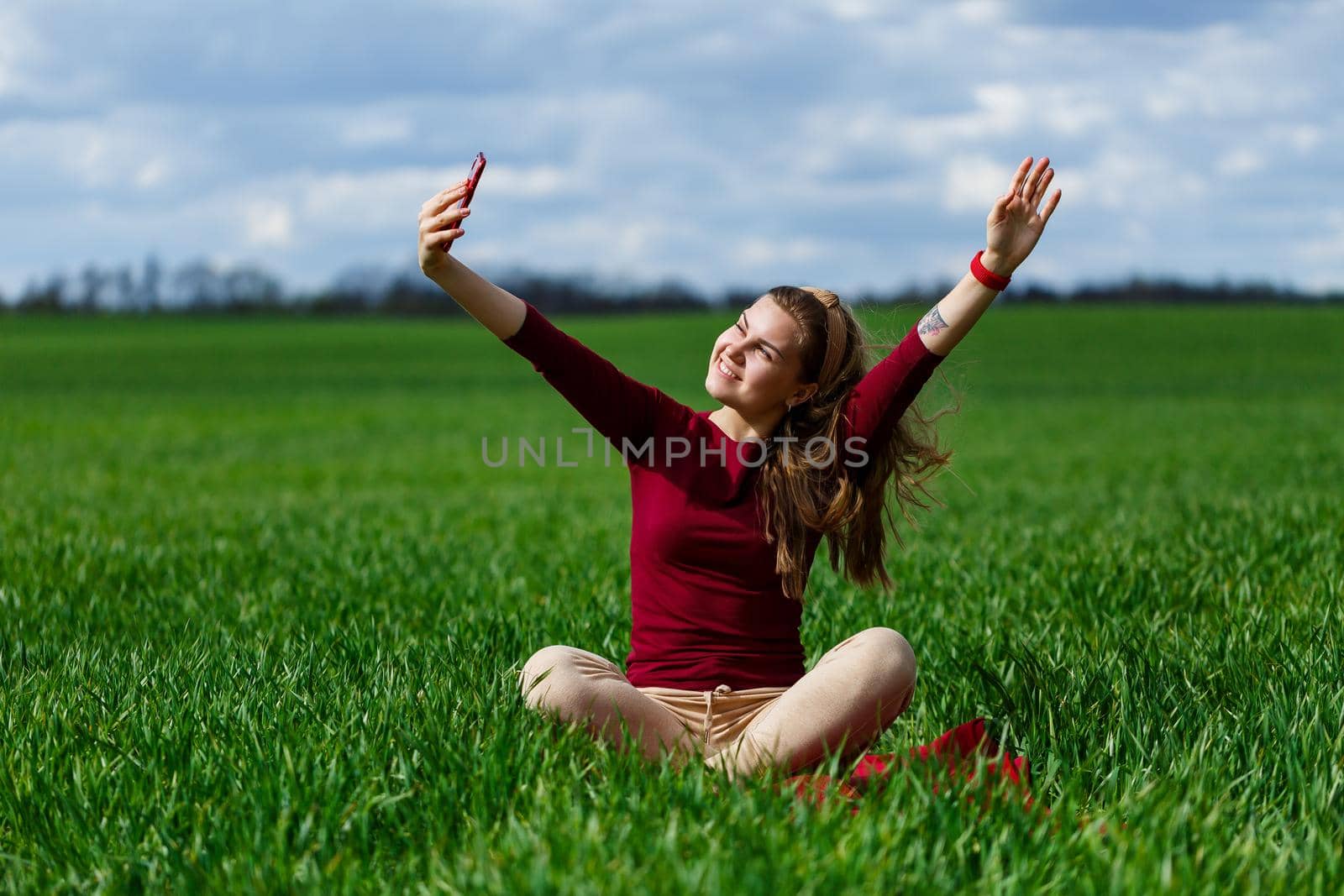 Young beautiful woman student with a phone in her hands sitting on the grass. Girl takes selfies and takes selfie pictures. She smiles and enjoys a warm day. Concept photo on smartphone