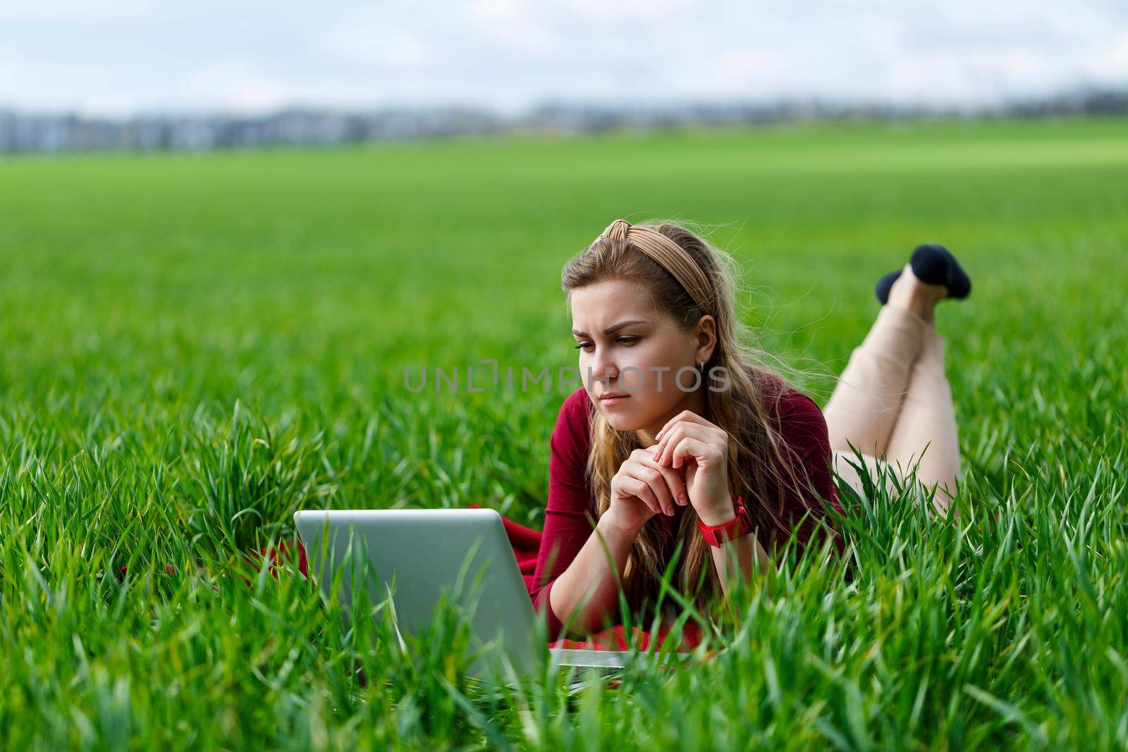 Beautiful young blonde woman is lying on the green grass in the park with a laptop and working. Blue sky with clouds. The girl smiles and enjoys a good day. Work on the nature on a sunny day.