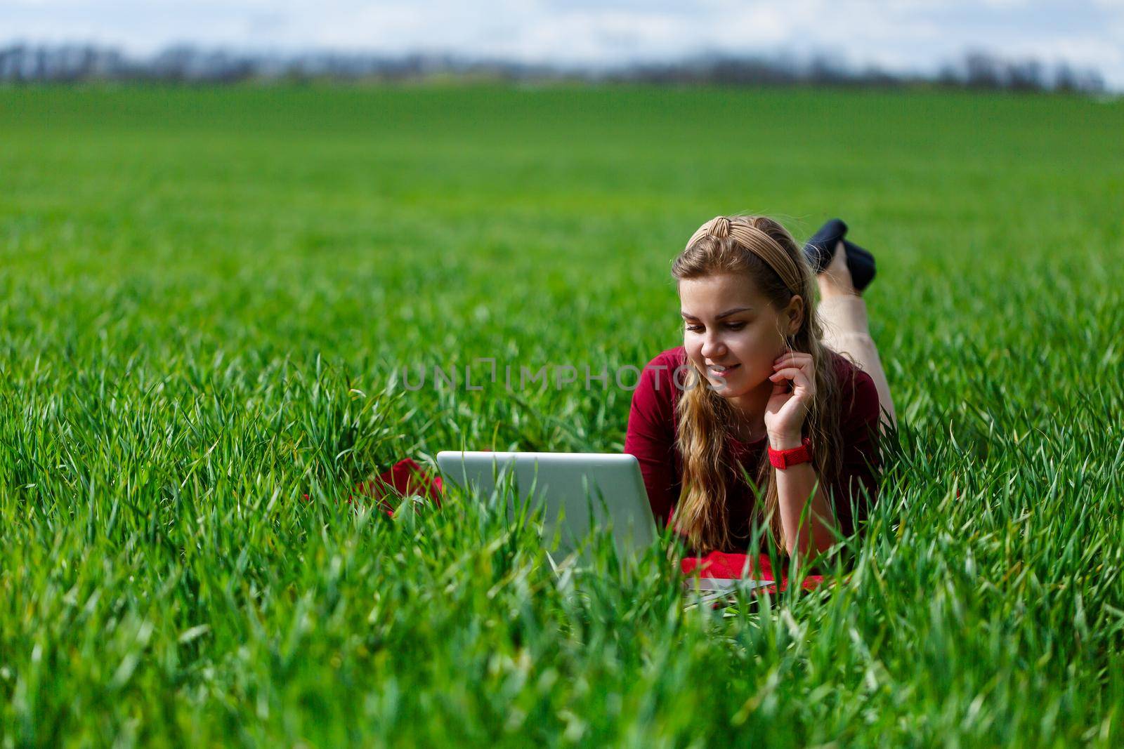 Beautiful young blonde woman is lying on the green grass in the park with a laptop and working. Blue sky with clouds. The girl smiles and enjoys a good day. Work on the nature on a sunny day.