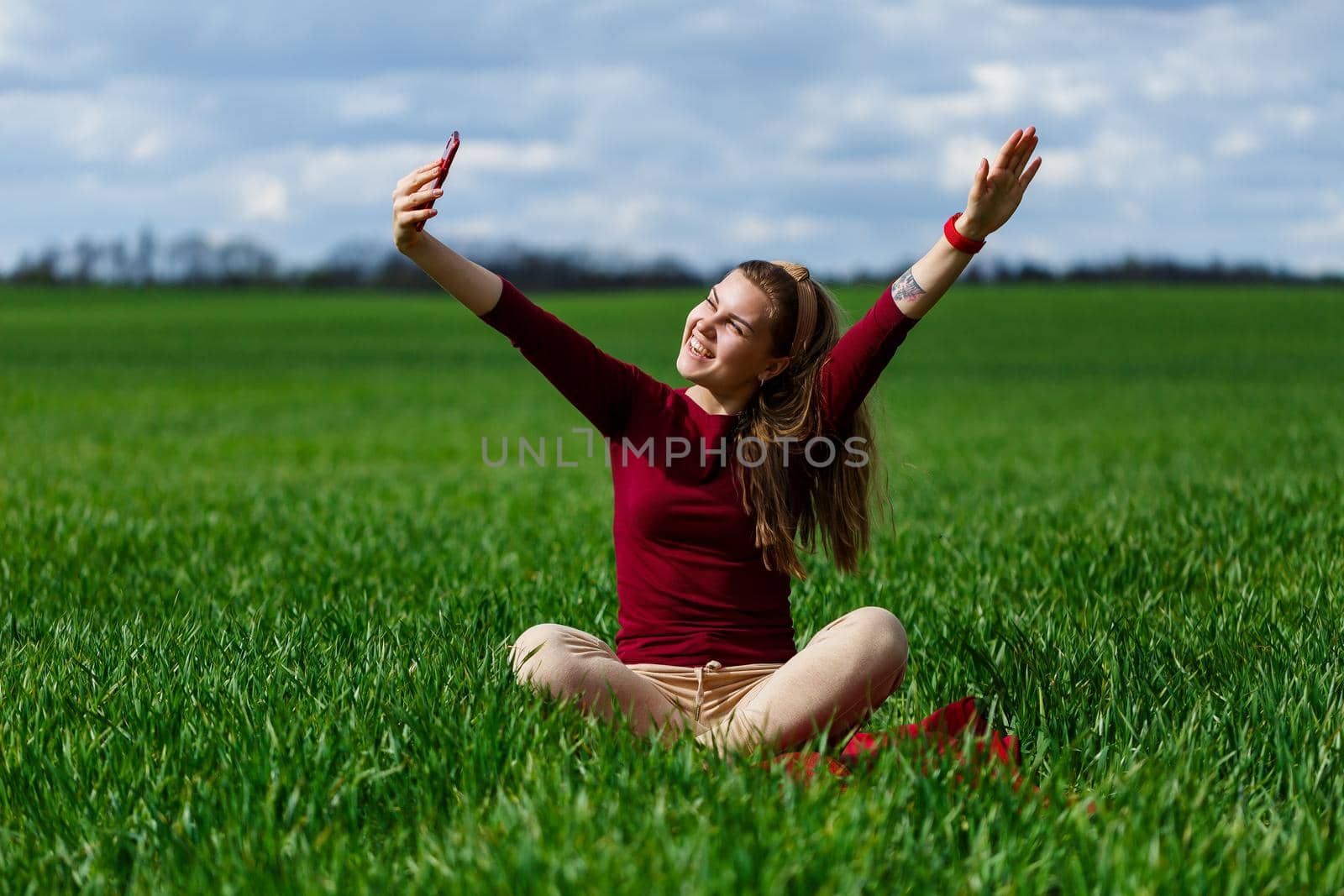 Young beautiful woman student with a phone in her hands sitting on the grass. Girl takes selfies and takes selfie pictures. She smiles and enjoys a warm day. Concept photo on smartphone