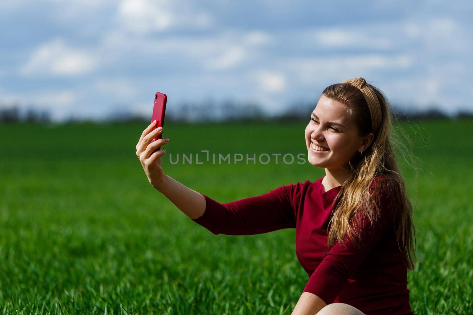 Beautiful woman with a phone in her hands sits on the grass. The girl looks at herself in the camera of the phone and takes selfie photos. She smiles and enjoys a warm day. Concept photo on smartphone
