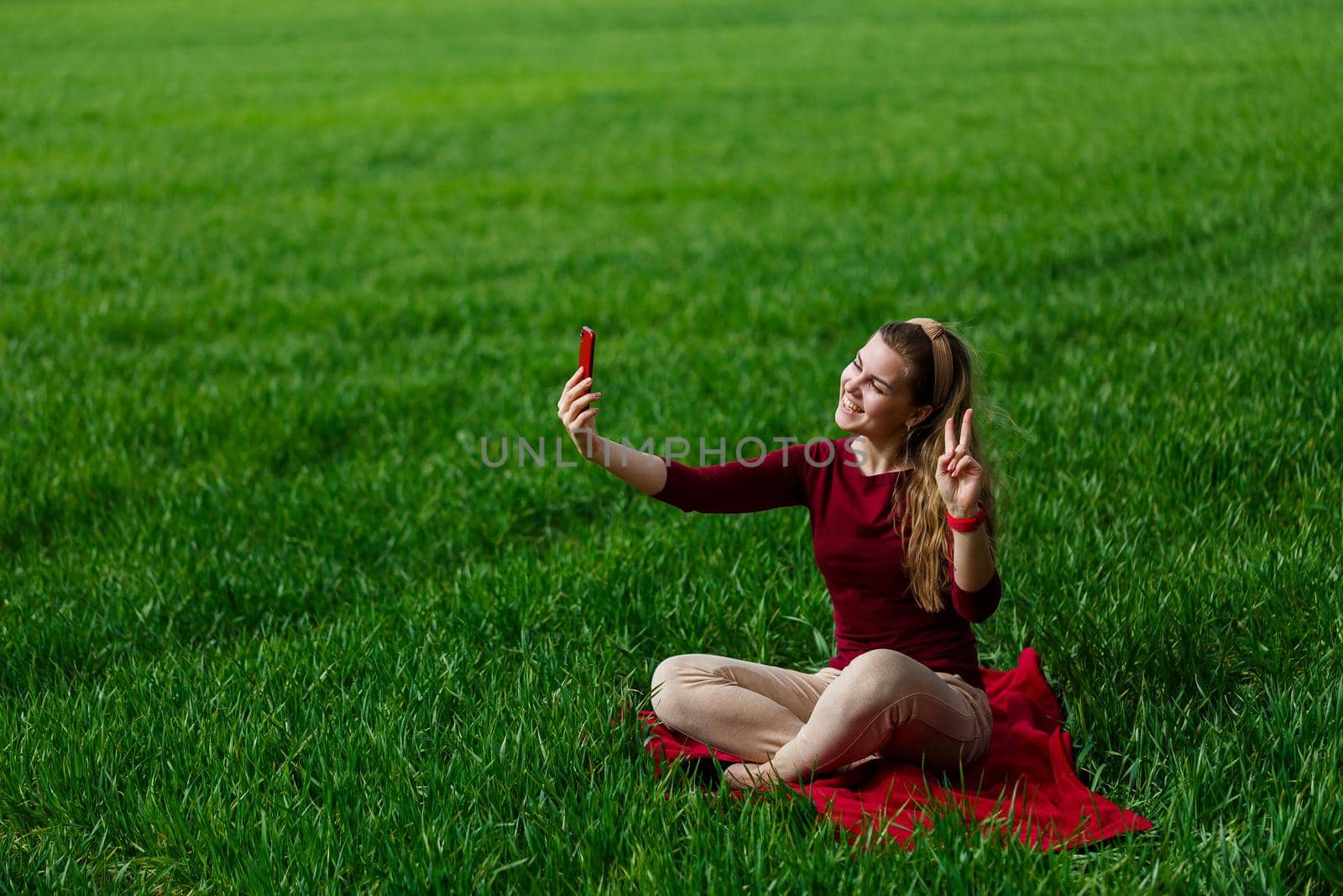 Beautiful woman with a phone in her hands sits on the grass. The girl looks at herself in the camera of the phone and takes selfie photos. She smiles and enjoys a warm day. Concept photo on smartphone