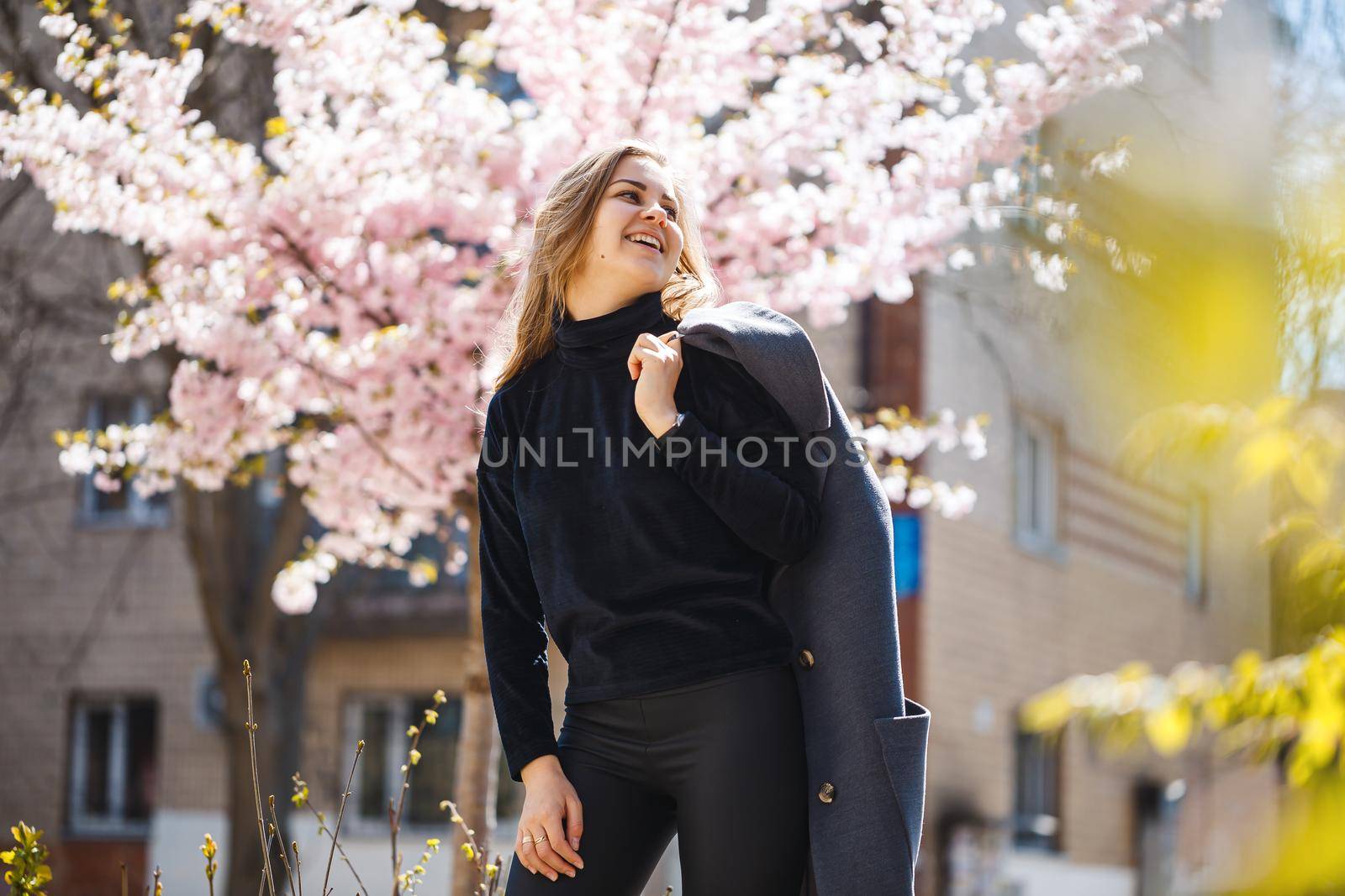 Sakura branches with flowers on a tree on the city streets. Happy woman girl in a gray palette walks along an alley with blooming sakura. Gorgeous fancy girl outdoors. Sakura tree blooming.