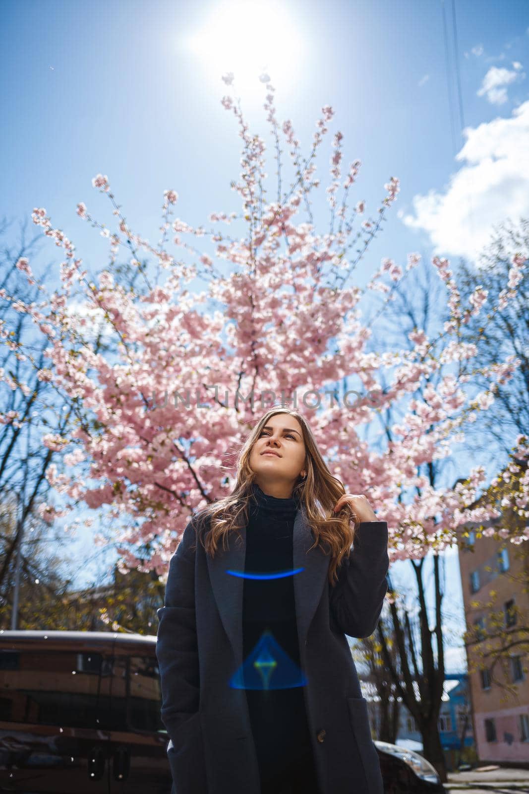 Sakura branches with flowers on a tree on the city streets. Stylish woman girl stands in the street with blooming sakura. Modern fashionable girl outdoors. Sakura tree blossoms.