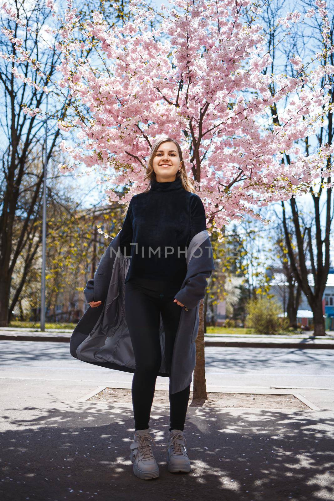 Young slender female model with long wavy hair and, dressed in a gray coat, sneakers, spinning on the street. Spring blossom trees woman girl laughs and runs to rejoice