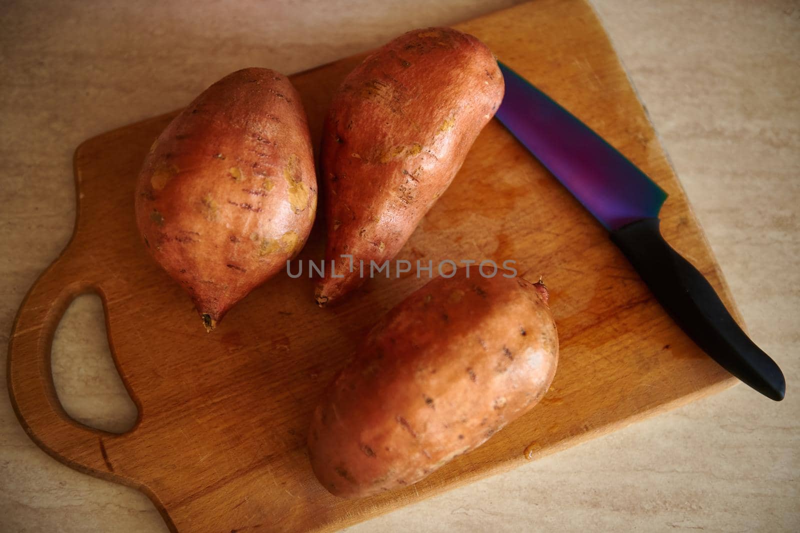 Top view of washed organic sweet potato tubers, batata and kitchen knife on a wooden cutting board on kitchen countertop by artgf