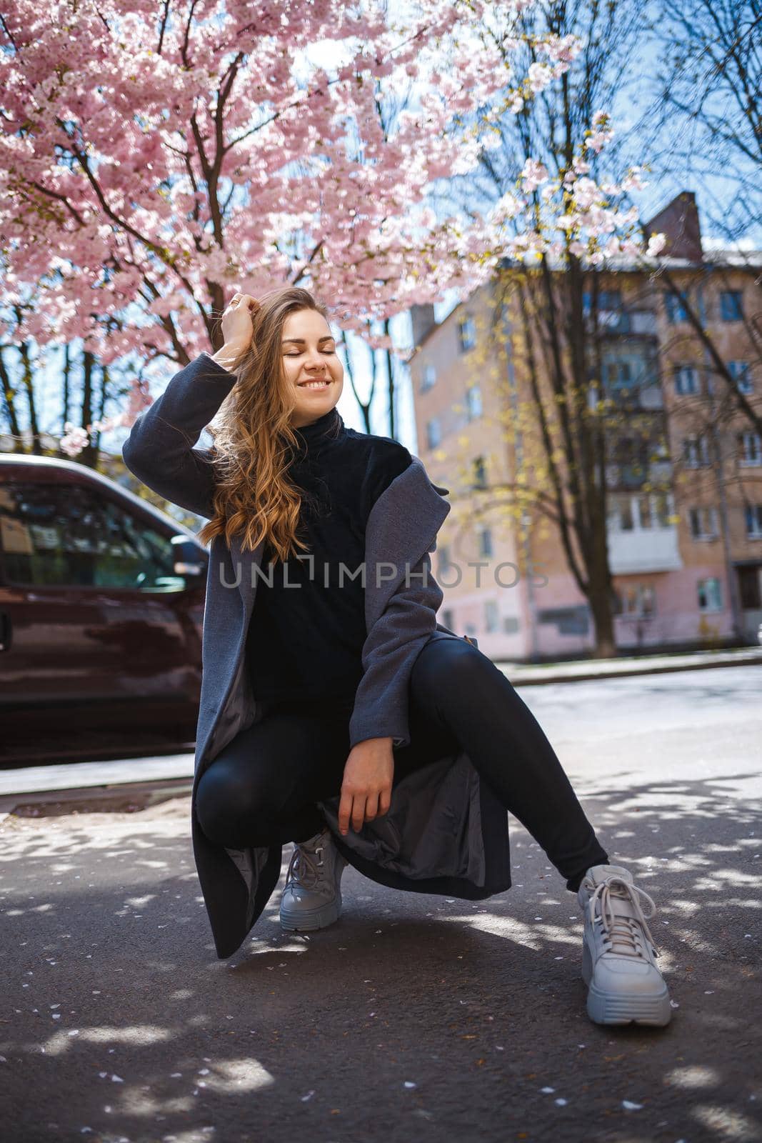 A young slender female model with long wavy hair and, dressed in a gray coat, sneakers, sits on the street. Spring blossom trees woman girl laugh and rejoice