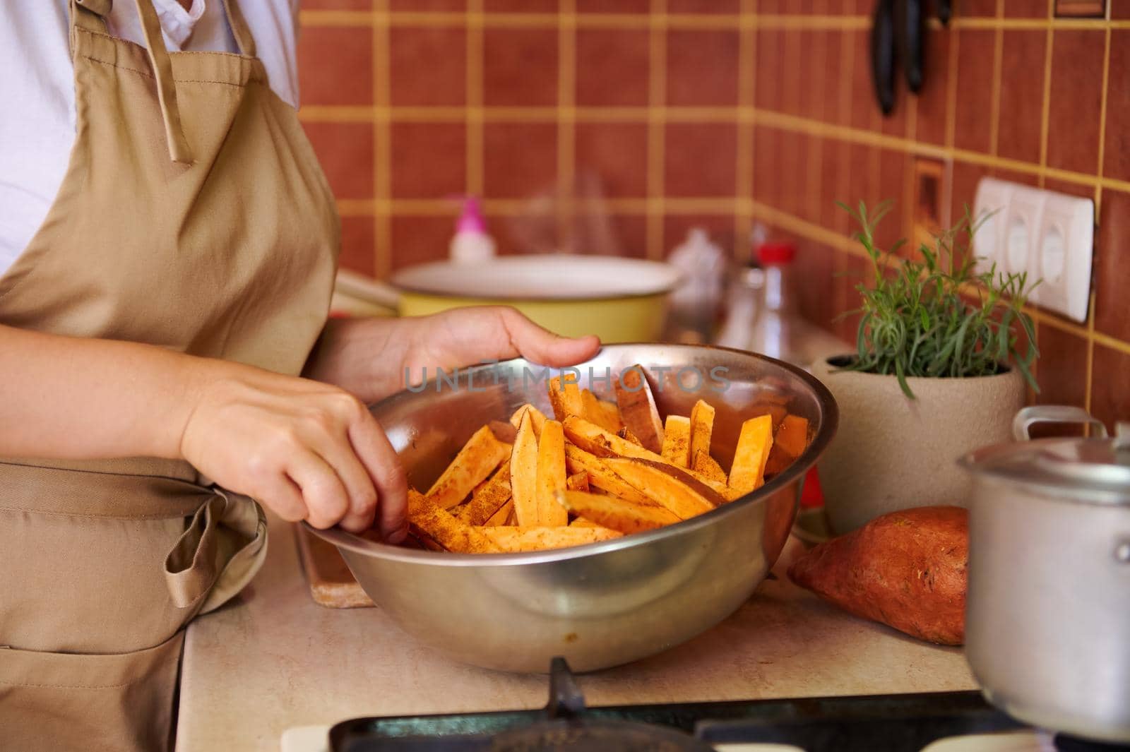 A stainless steel metal bowl with slices of sweet potatoes, seasoned with fragrant culinary herbs, in the hands of a housewife in chef's apron mixing ingredients, while cooking delicious healthy dish