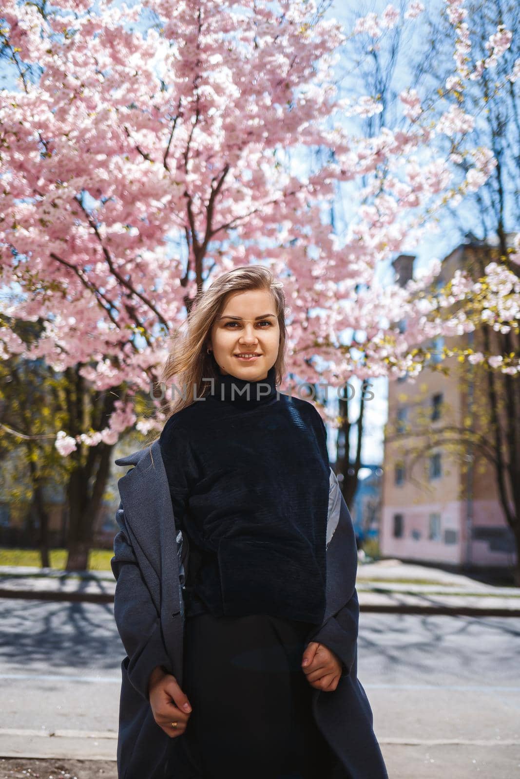 A young slender female model with long wavy hair and, dressed in a gray coat, sneakers, stands on the street near a flowering shrub with beautiful pink flowers in the background and poses.