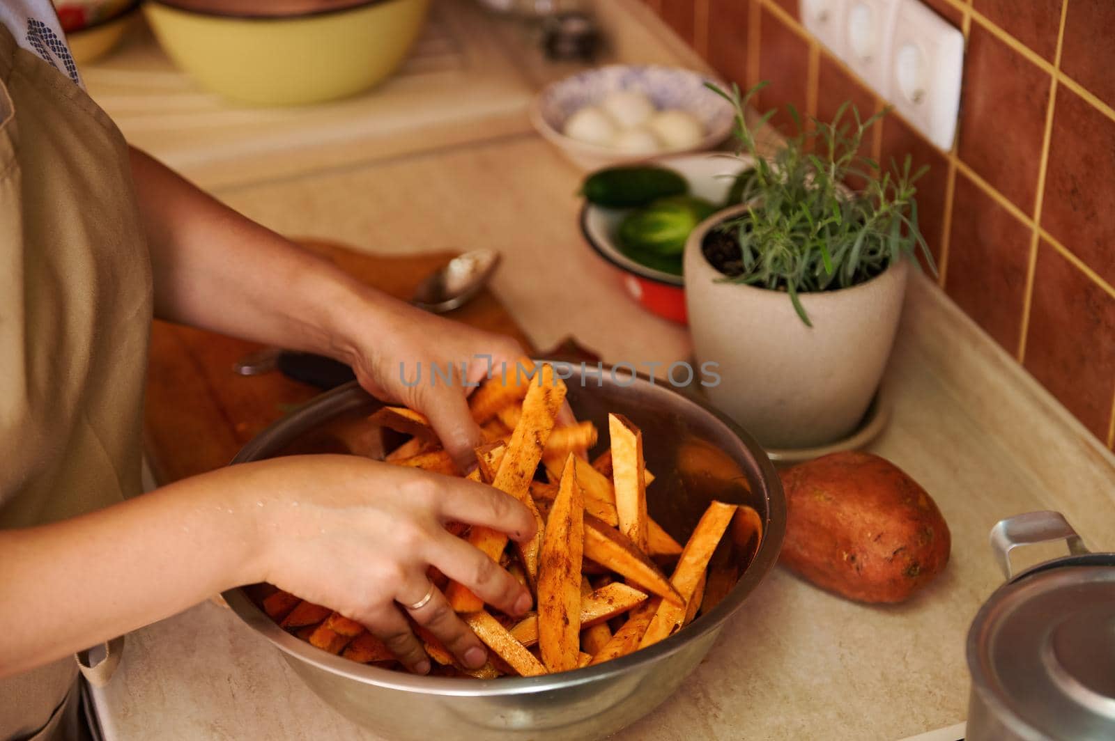 Close-up housewife standing by kitchen table and seasoning sliced wedges of organic batata with fragrant culinary herbs by artgf