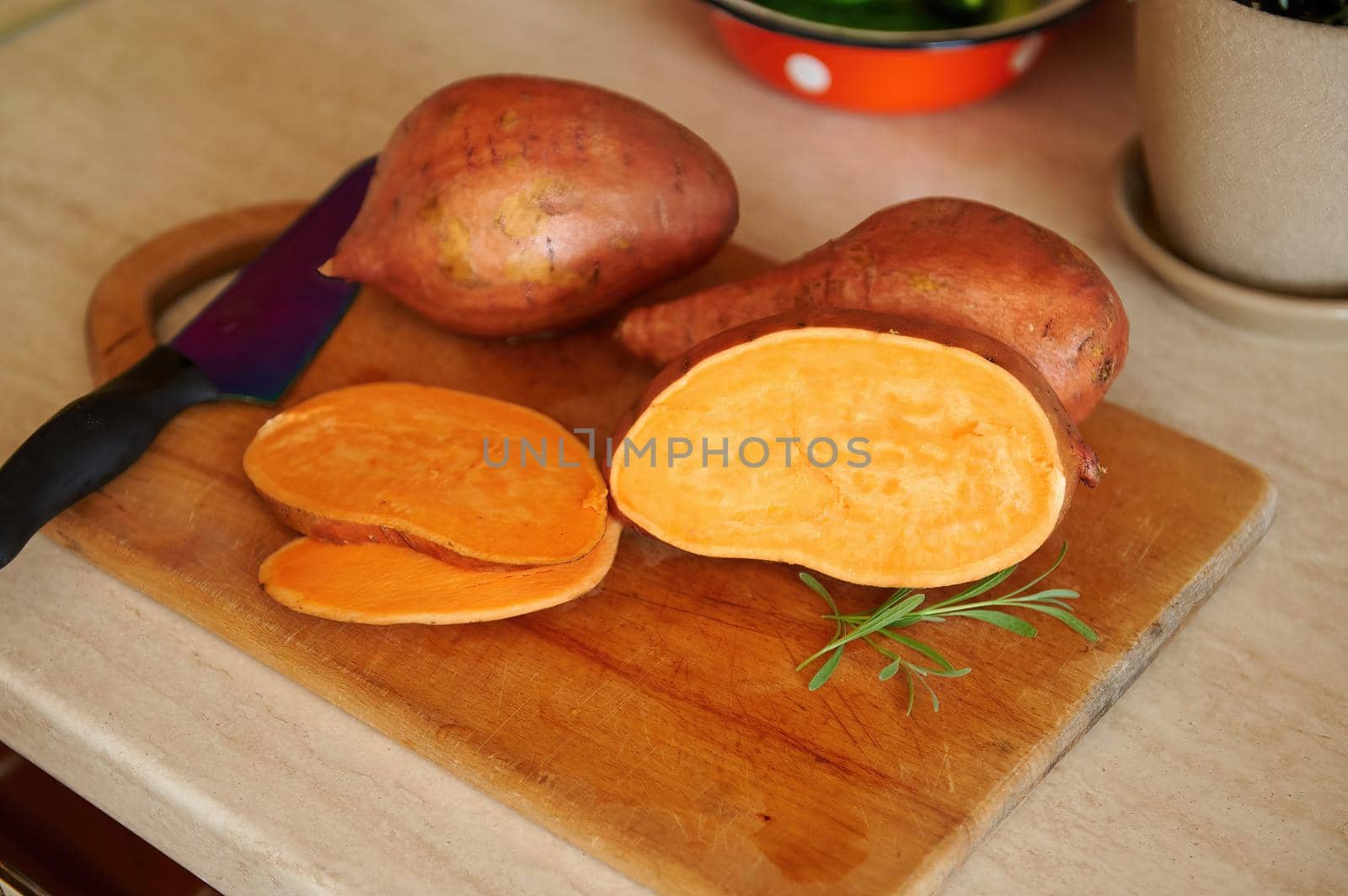 View from above of a whole and sliced tuber of organic raw orange color sweet potato, or batata, a kitchen knife and rosemary leaves for seasoning lying down on a wooden cutting board.