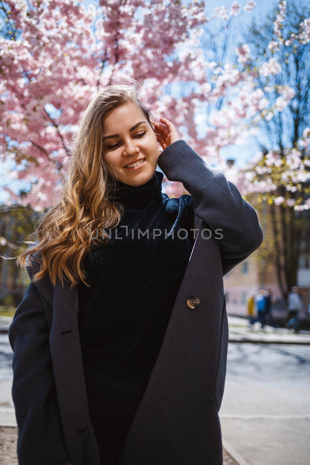 A young slender female model with long wavy hair and, dressed in a gray coat, sneakers, stands on the street near a flowering shrub with beautiful pink flowers in the background and poses.