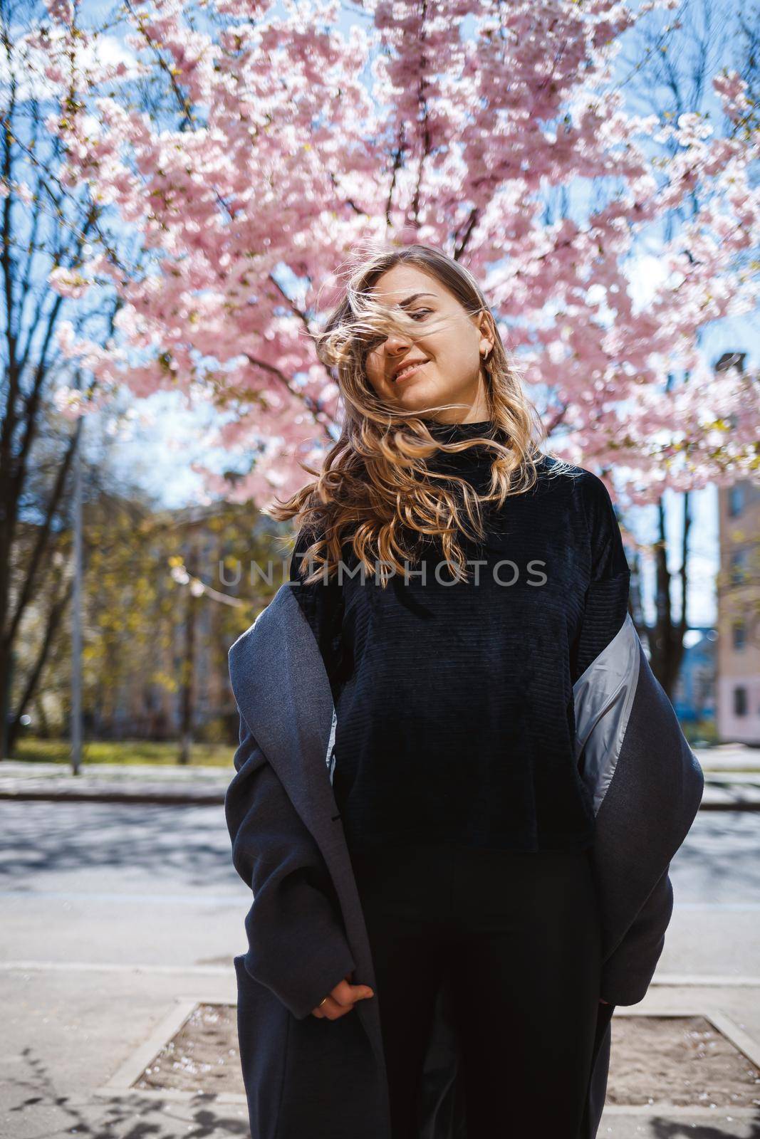 Young slender female model with long wavy hair and, dressed in a gray coat, sneakers, spinning on the street. Spring blossom trees woman girl laughs and runs to rejoice