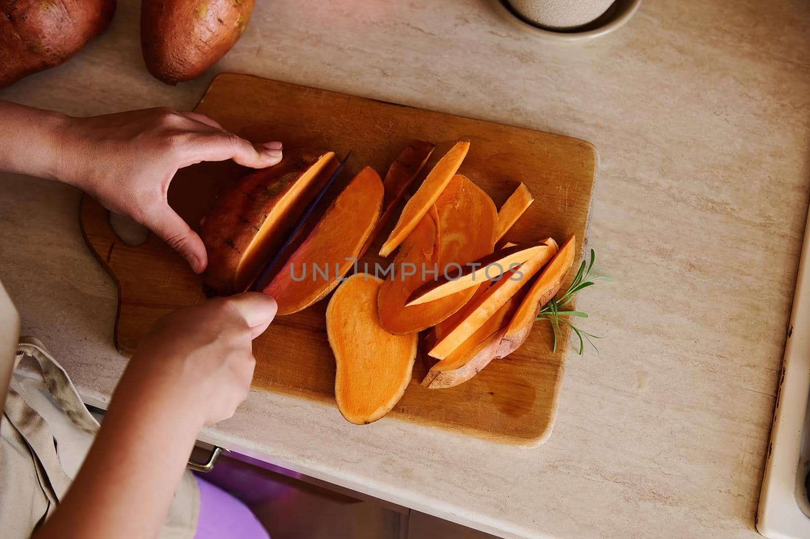 Overhead view of a woman, a housewife cutting a whole tuber of sweet potato into slices on a wooden board in the kitchen by artgf