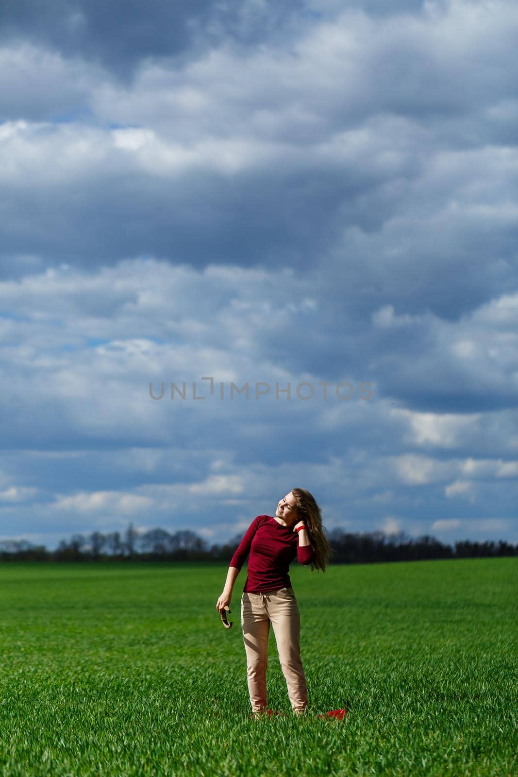 Beautiful young blonde woman stands on green grass in a park. Blue sky with clouds. The girl smiles and enjoys a good day.