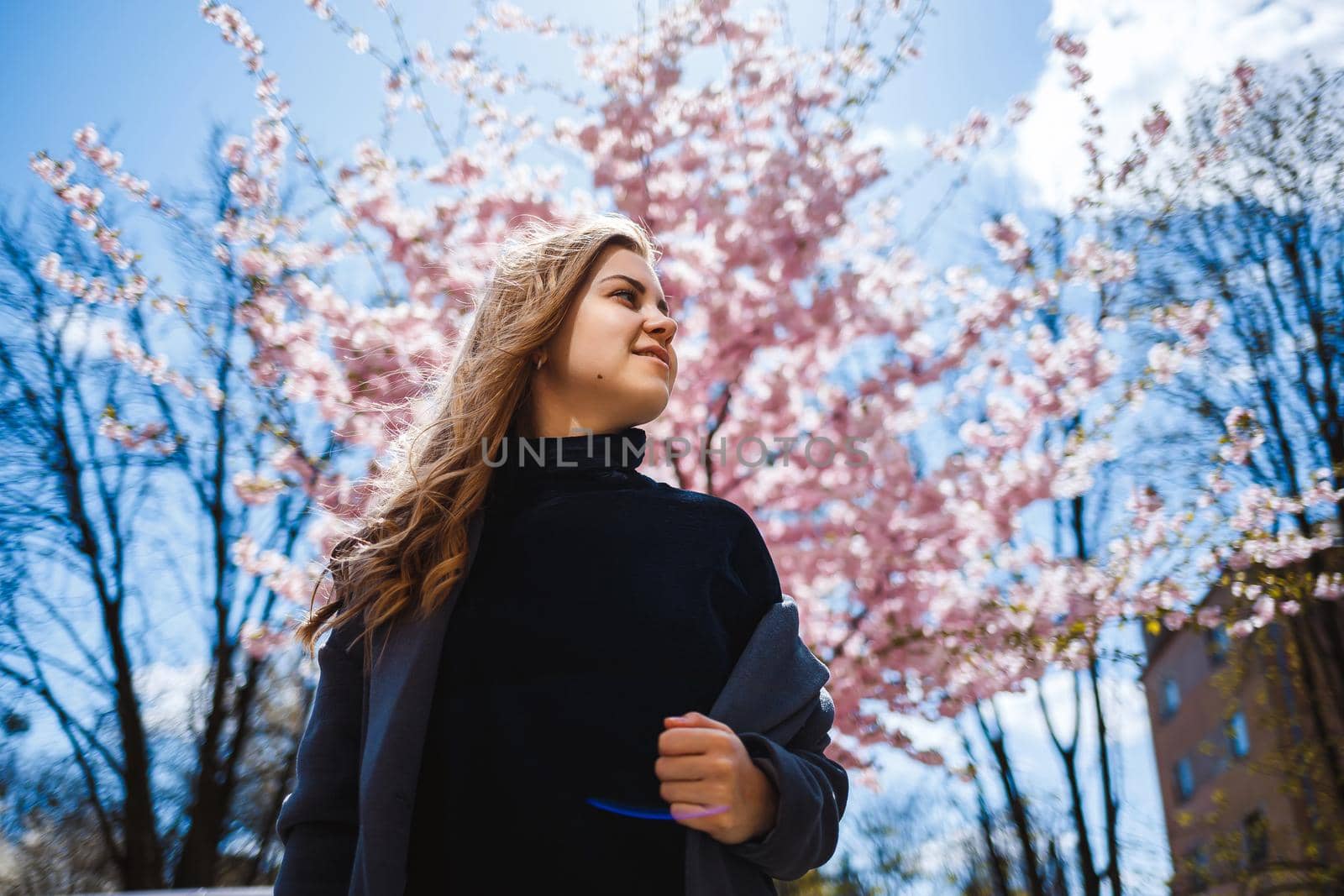 Sakura branches with flowers on a tree on the city streets. Stylish woman girl stands in the street with blooming sakura. Modern fashionable girl outdoors. Sakura tree blossoms.