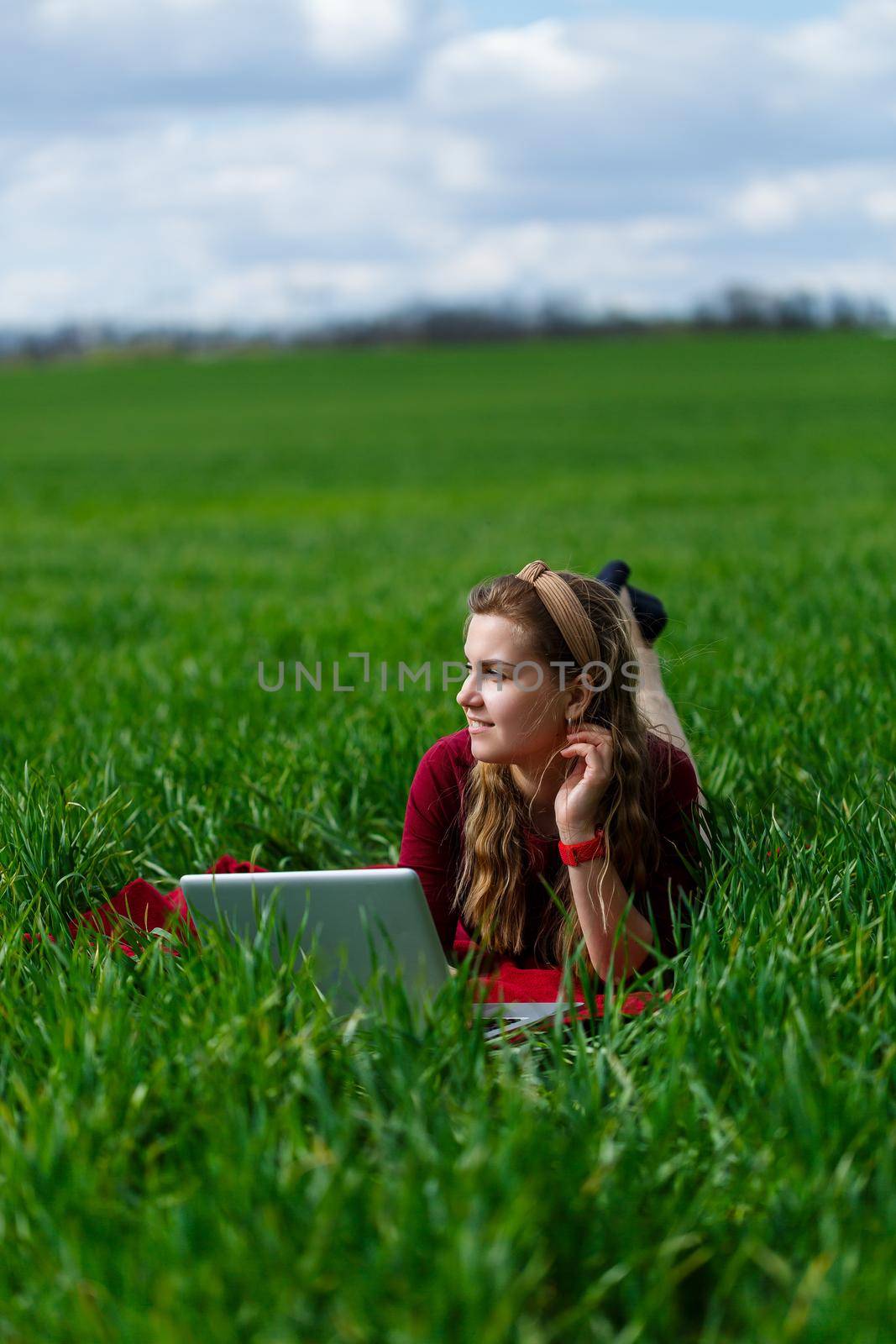 Beautiful young blonde woman is lying on the green grass in the park with a laptop and working. Blue sky with clouds. The girl smiles and enjoys a good day. Work on the nature on a sunny day. by Dmitrytph