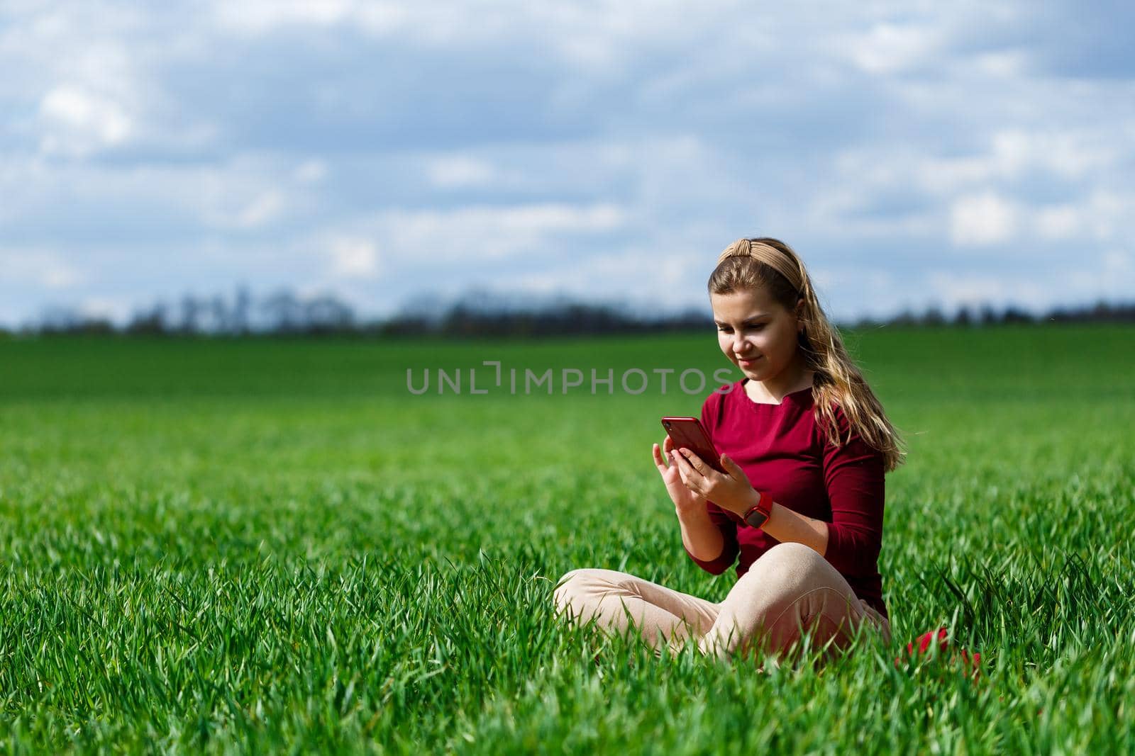 Young beautiful woman student with a phone in her hands sitting on the grass. Girl takes selfies and takes selfie pictures. She smiles and enjoys a warm day. Concept photo on smartphone