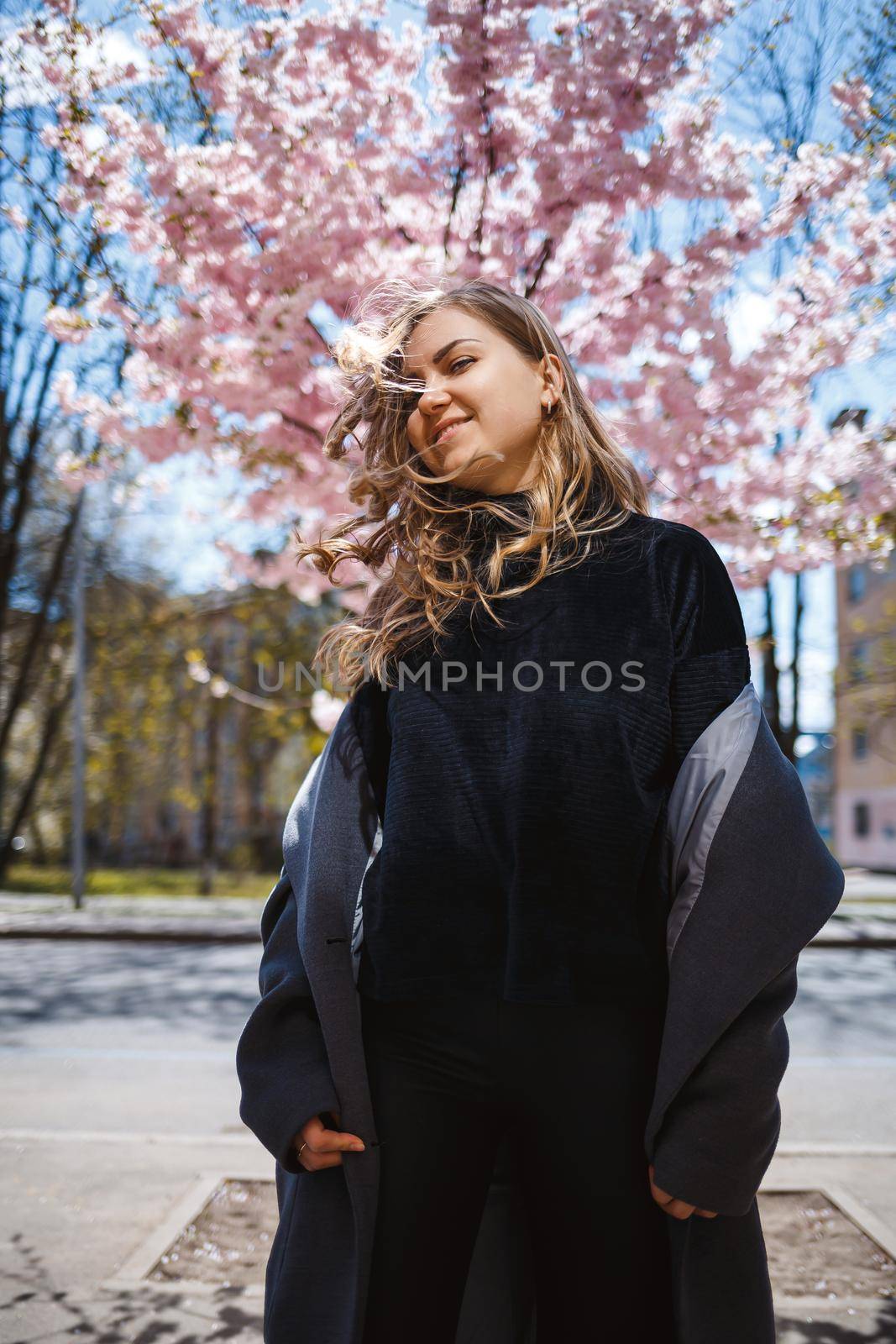 Young slender female model with long wavy hair and, dressed in a gray coat, sneakers, spinning on the street. Spring blossom trees woman girl laughs and runs to rejoice