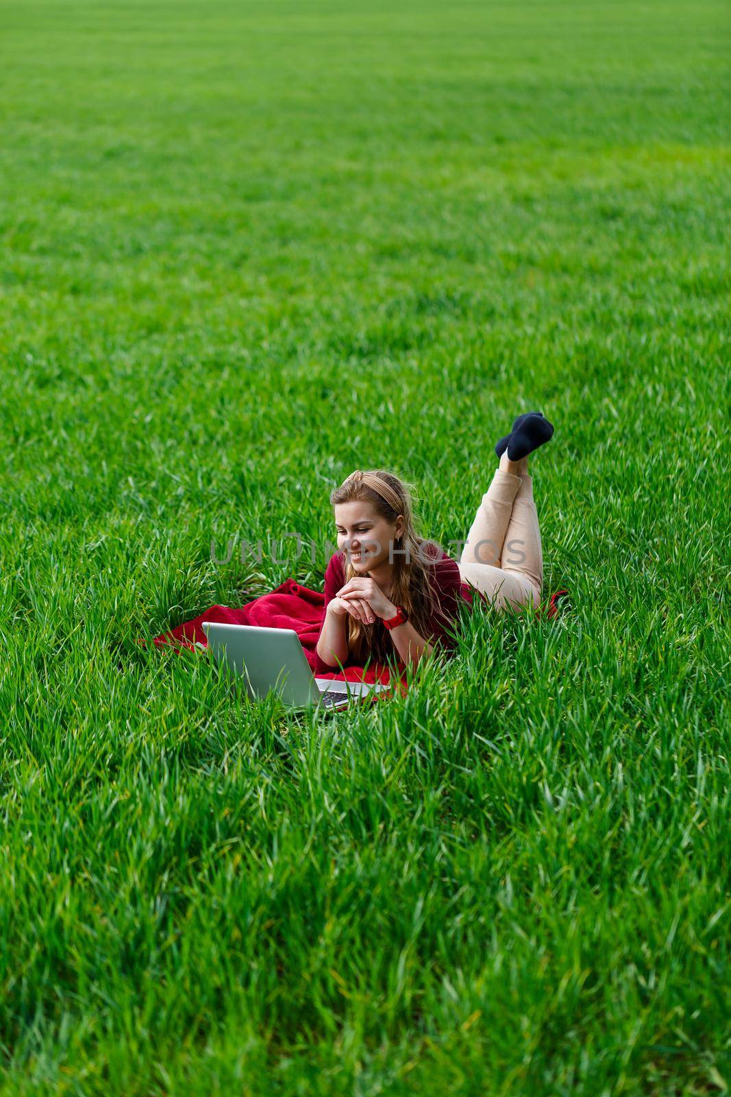 Beautiful young blonde woman is lying on the green grass in the park with a laptop and working. Blue sky with clouds. The girl smiles and enjoys a good day. Work on the nature on a sunny day.