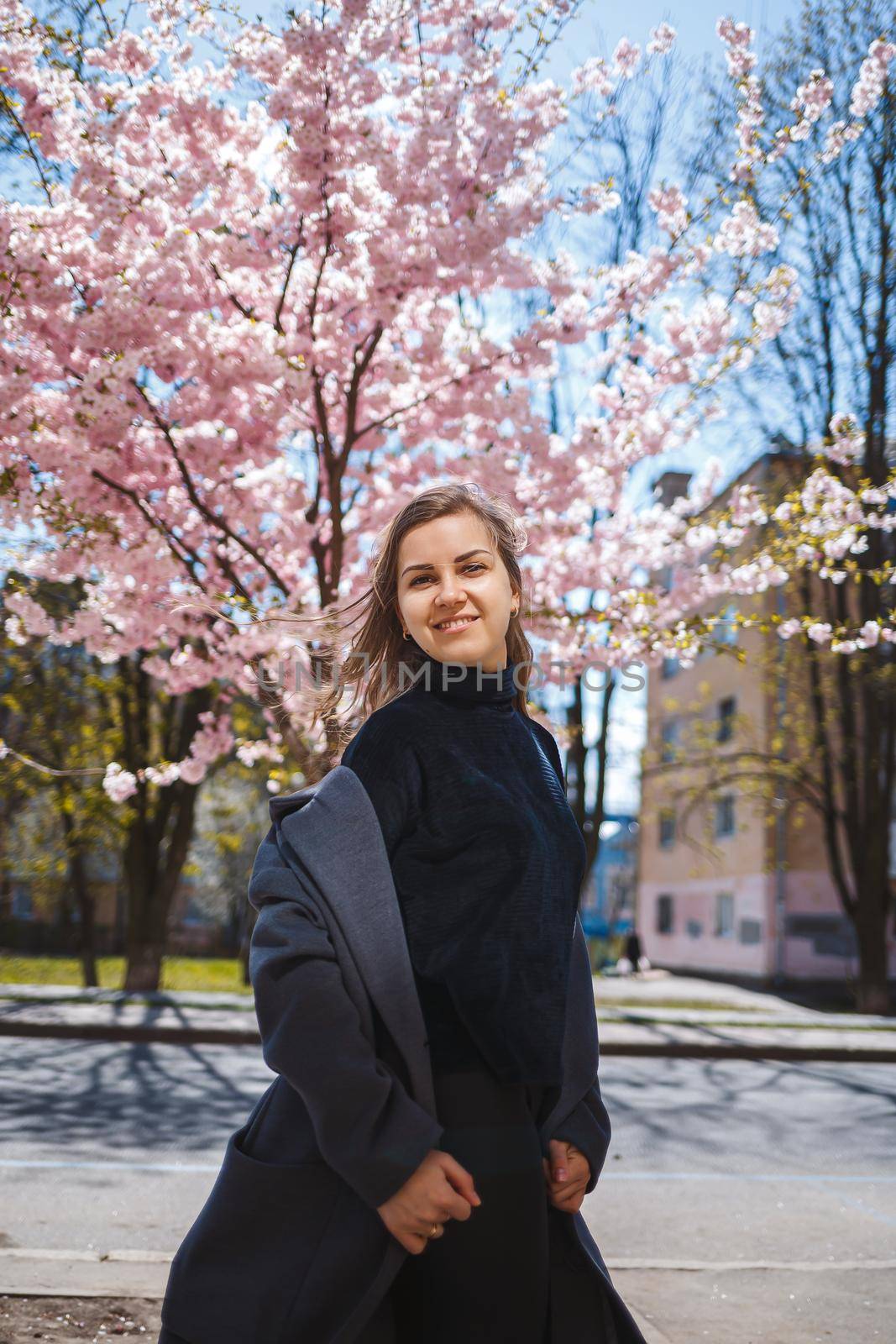Young slender female model with long wavy hair and, dressed in a gray coat, sneakers, spinning on the street. Spring blossom trees woman girl laughs and runs to rejoice