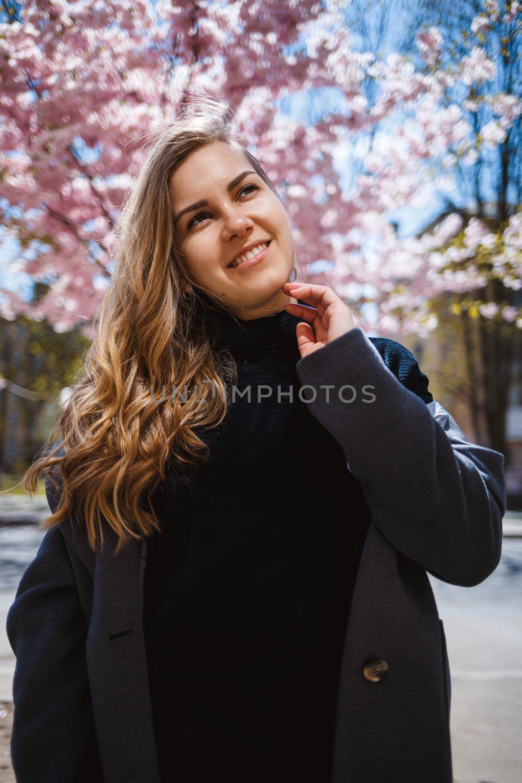 A young slender female model with long wavy hair and, dressed in a gray coat, sneakers, stands on the street near a flowering shrub with beautiful pink flowers in the background and poses.
