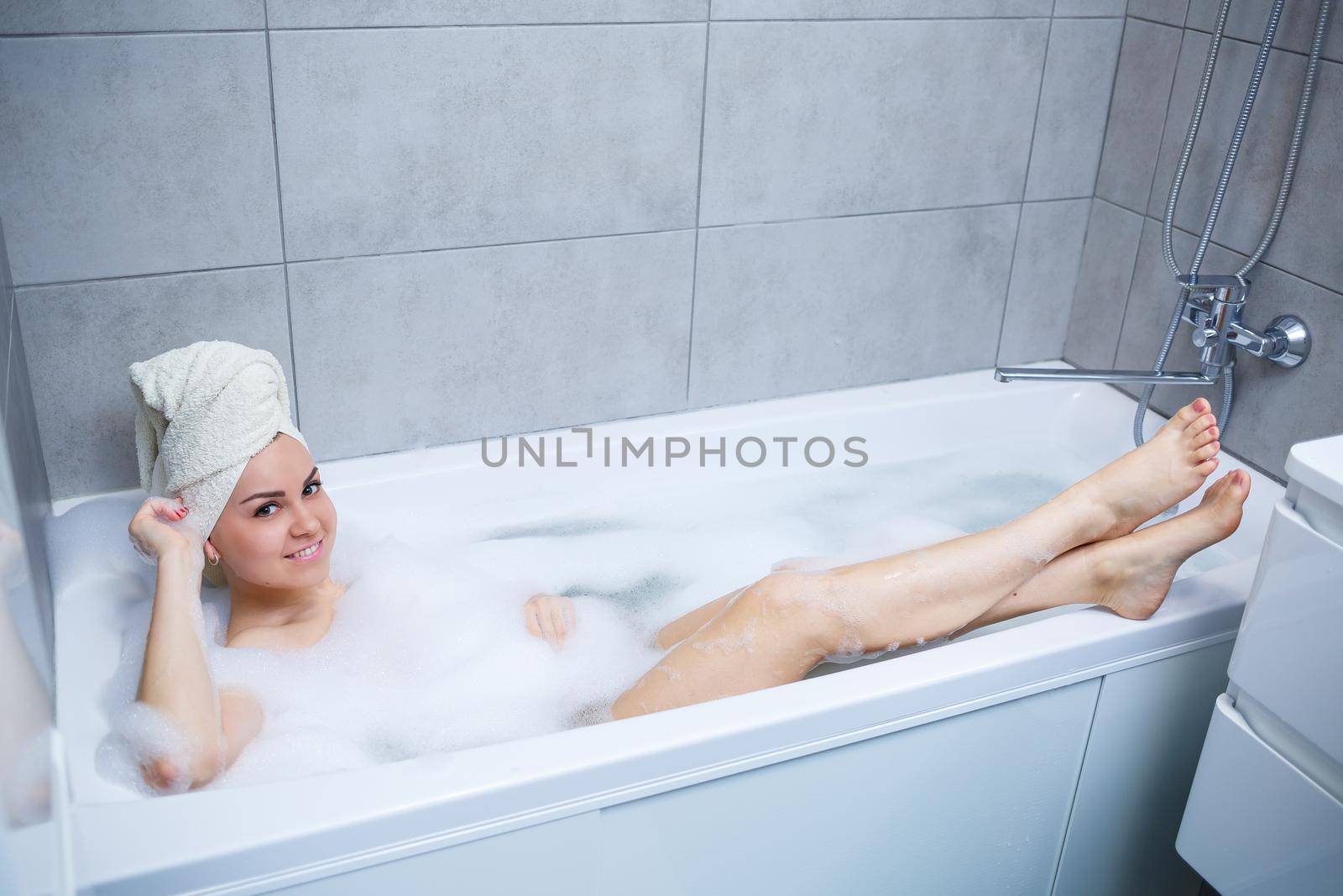 Girl woman with a white towel on her head lies in the bathroom in a white bath. In the hands of a lot of muddy soap. Full whirlpool foam. Relax after a hard day. Spa relaxing treatment