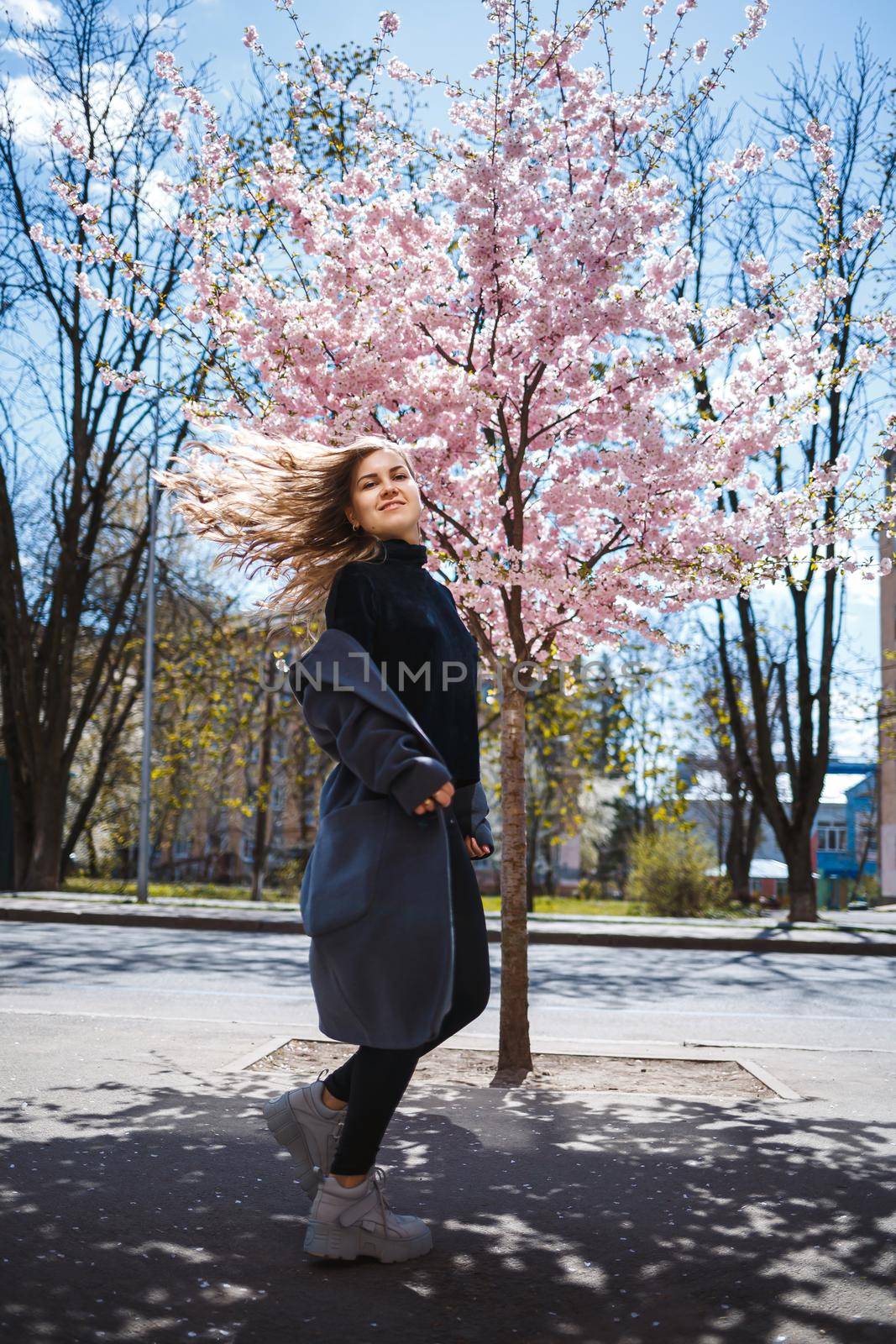 Young slender female model with long wavy hair and, dressed in a gray coat, sneakers, spinning on the street. Spring blossom trees woman girl laughs and runs to rejoice