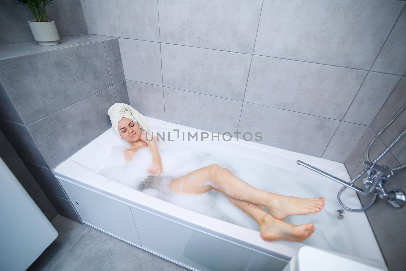 Girl woman with a white towel on her head lies in the bathroom in a white bath. In the hands of a lot of muddy soap. Full whirlpool foam. Relax after a hard day. Spa relaxing treatment