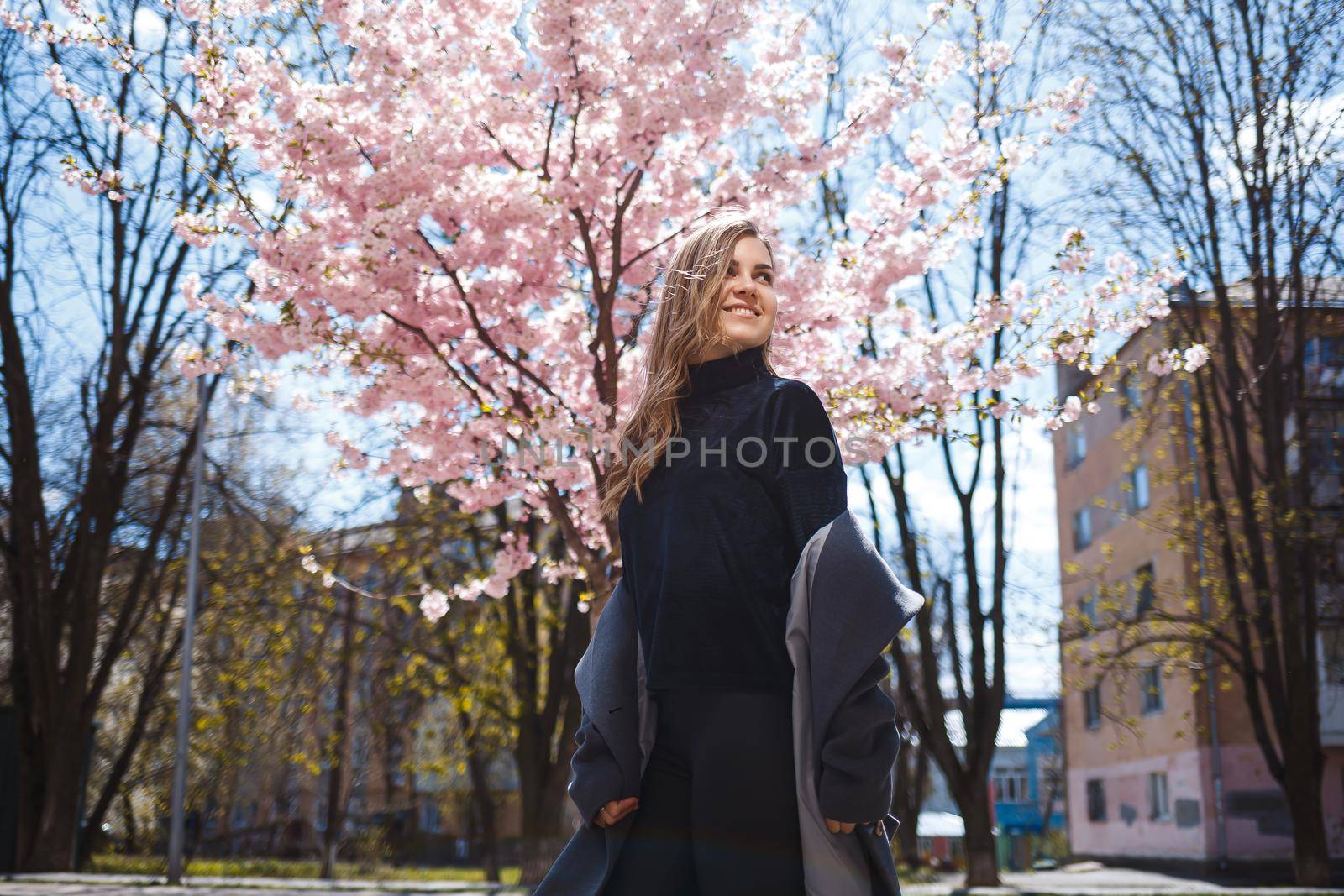 A young slender female model with long wavy hair and, dressed in a gray coat, sneakers, stands on the street near a flowering shrub with beautiful pink flowers in the background and poses.