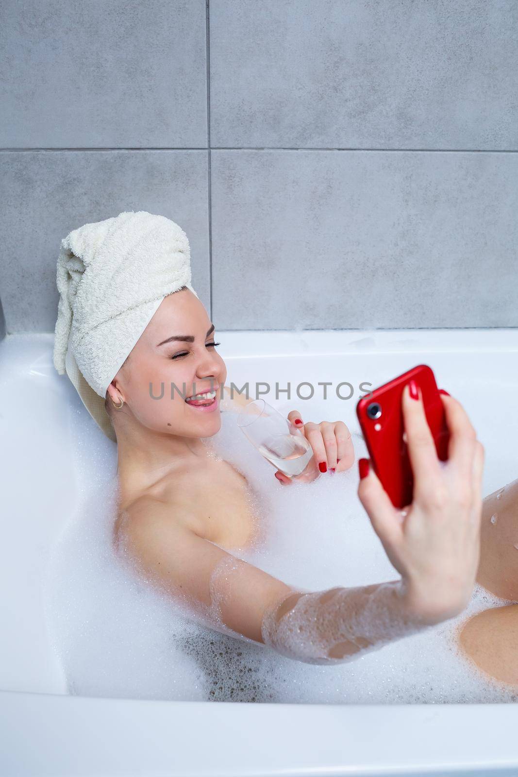 Girl woman with a towel on her head lies with a glass of champagne in a white bathtub and takes a selfie on the phone. In her hands a lot of drunk soap. Relax after a hard day. Spa relaxing procedure