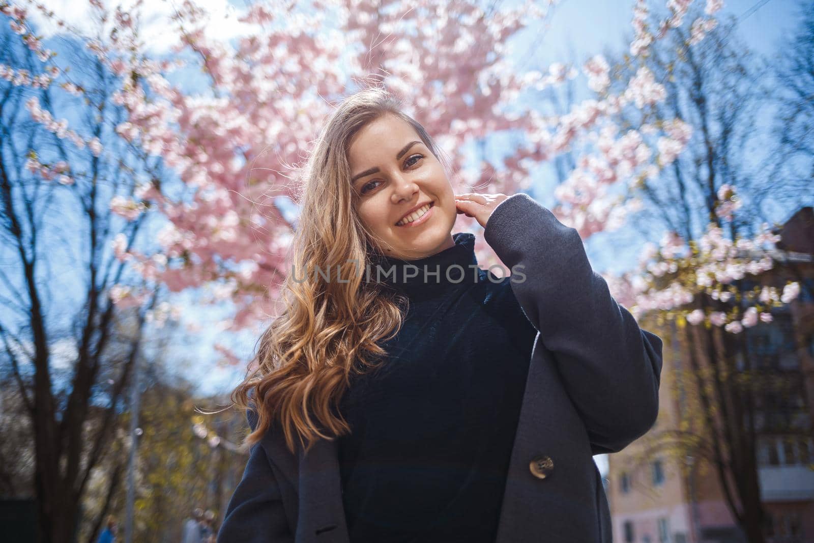 A young slender female model with long wavy hair and, dressed in a gray coat, sneakers, stands on the street near a flowering shrub with beautiful pink flowers in the background and poses.