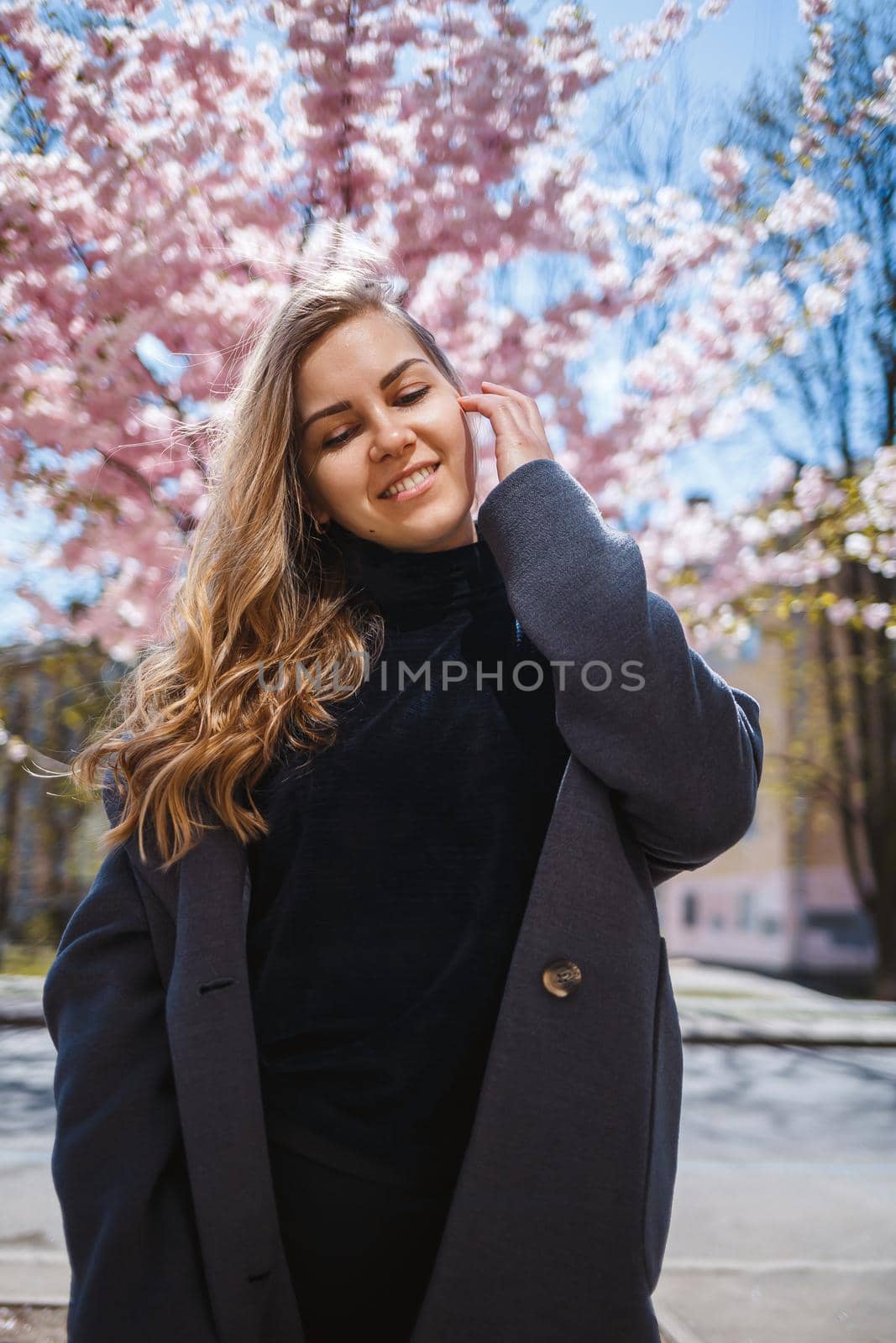 A young slender female model with long wavy hair and, dressed in a gray coat, sneakers, stands on the street near a flowering shrub with beautiful pink flowers in the background and poses.