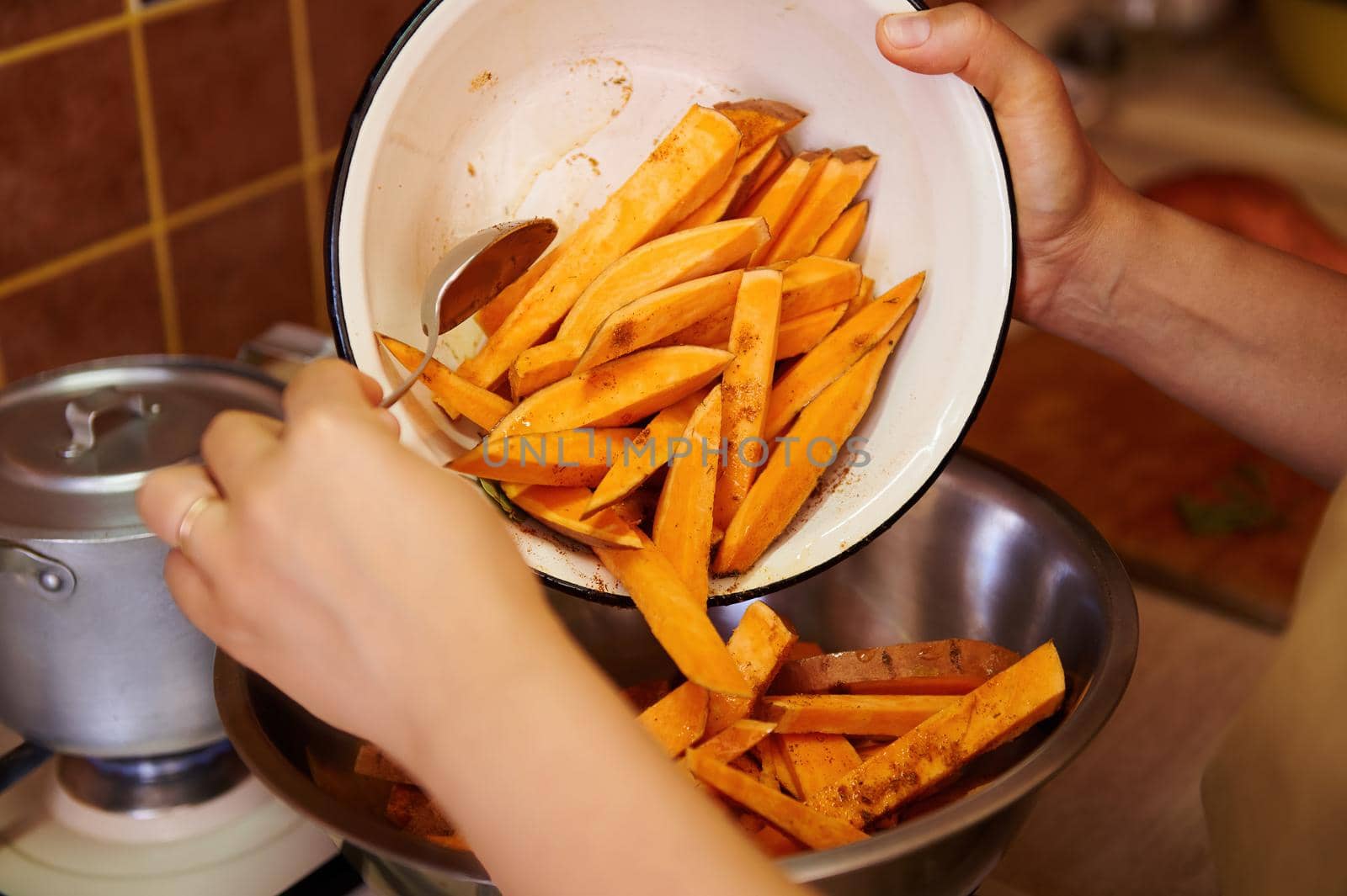 Selective focus on wedges of organic sweet potato in an enamel bowl, in the hands of a cook seasoning slices batata and mixing ingredients while preparing healthy vegan meal in the home kitchen