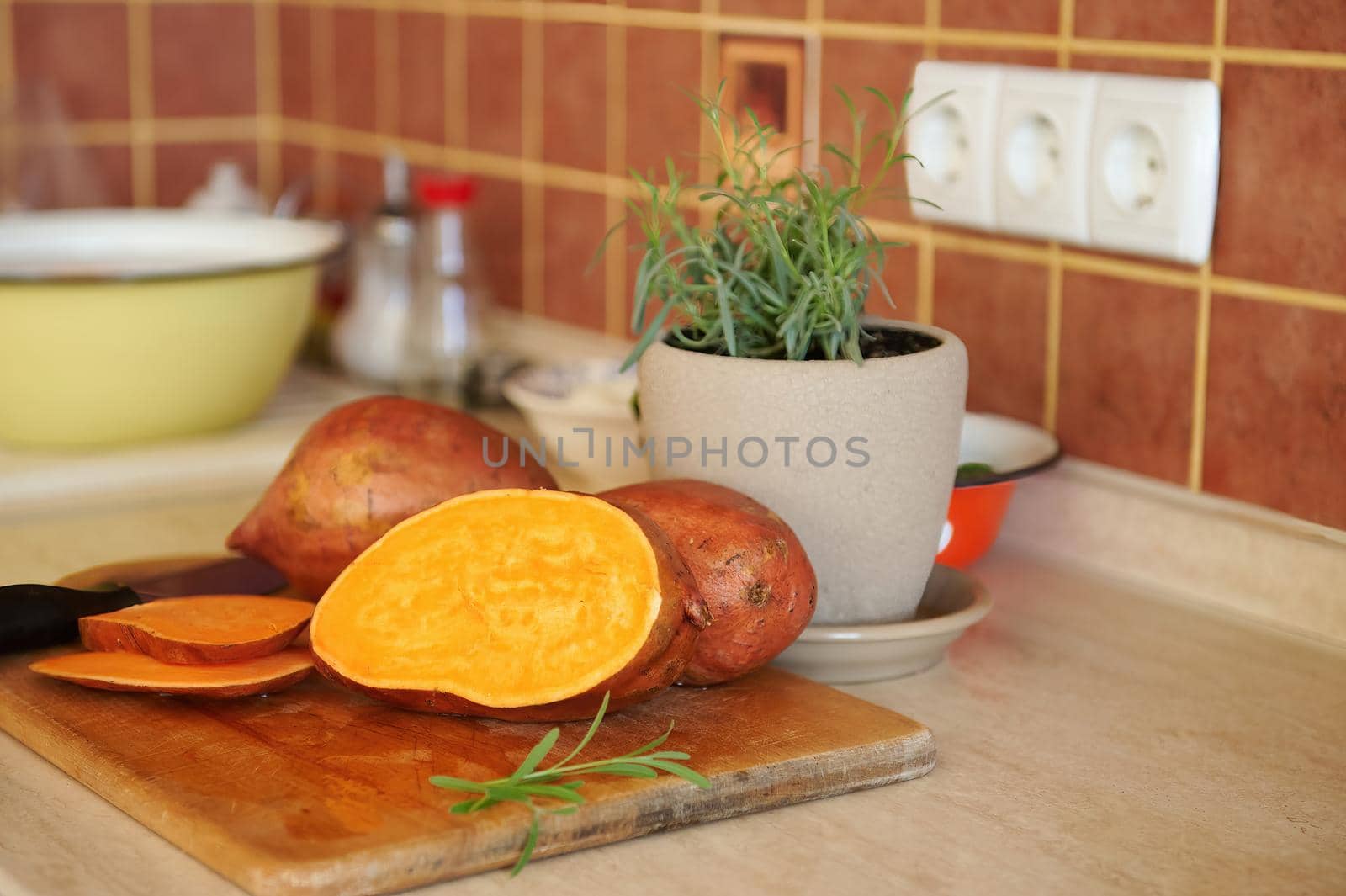 Close-up of whole and sliced tuber of sweet potato, batata, a kitchen knife on wooden board and growing rosemary in pot by artgf