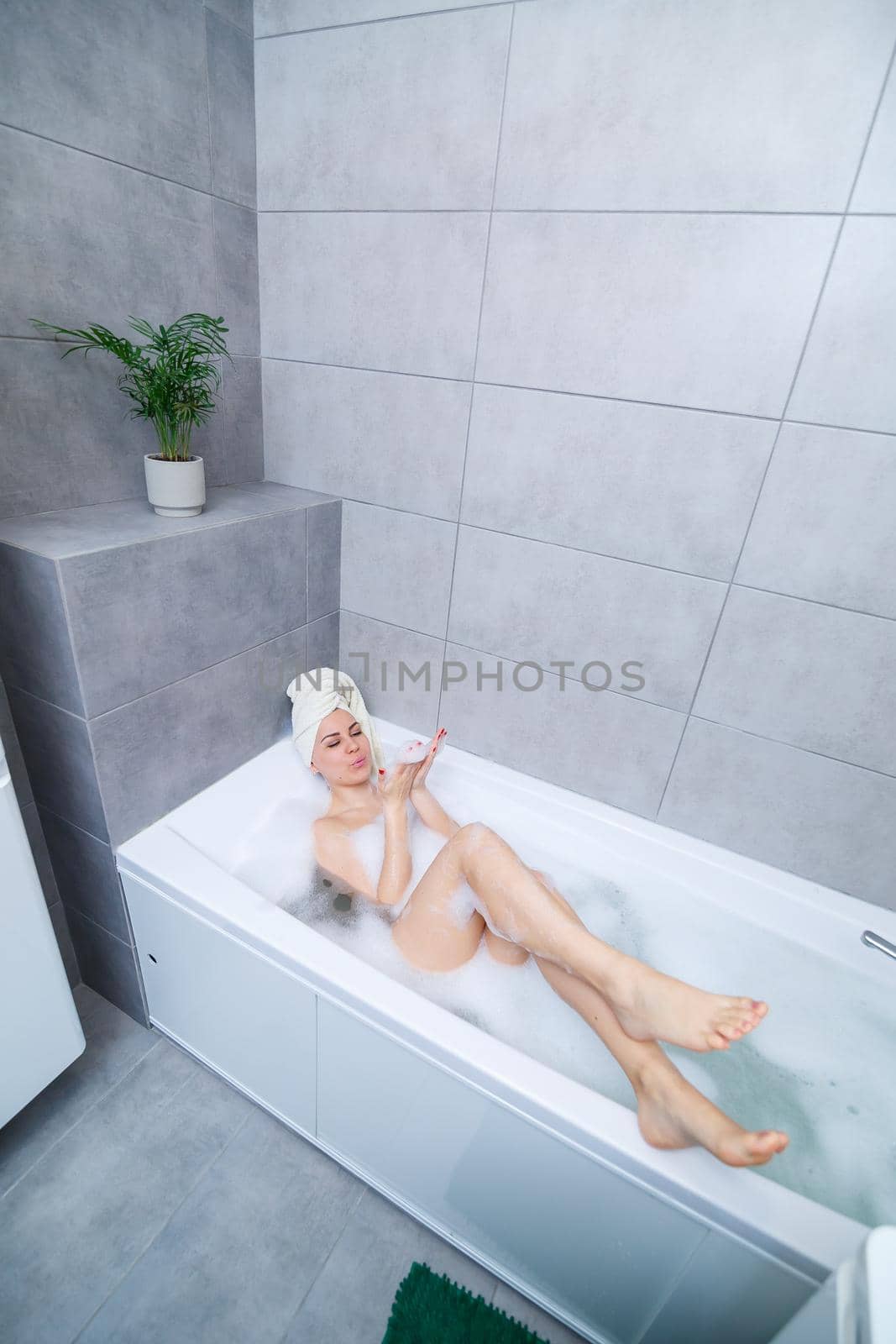 Girl woman with a white towel on her head lies in the bathroom in a white bath. In the hands of a lot of muddy soap. Full whirlpool foam. Relax after a hard day. Spa relaxing treatment