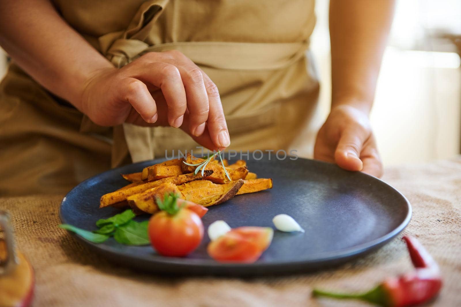 Close-up housewife in chef apron decorating dishes of fried sweet potato with cherry tomatoes and culinary herbs by artgf