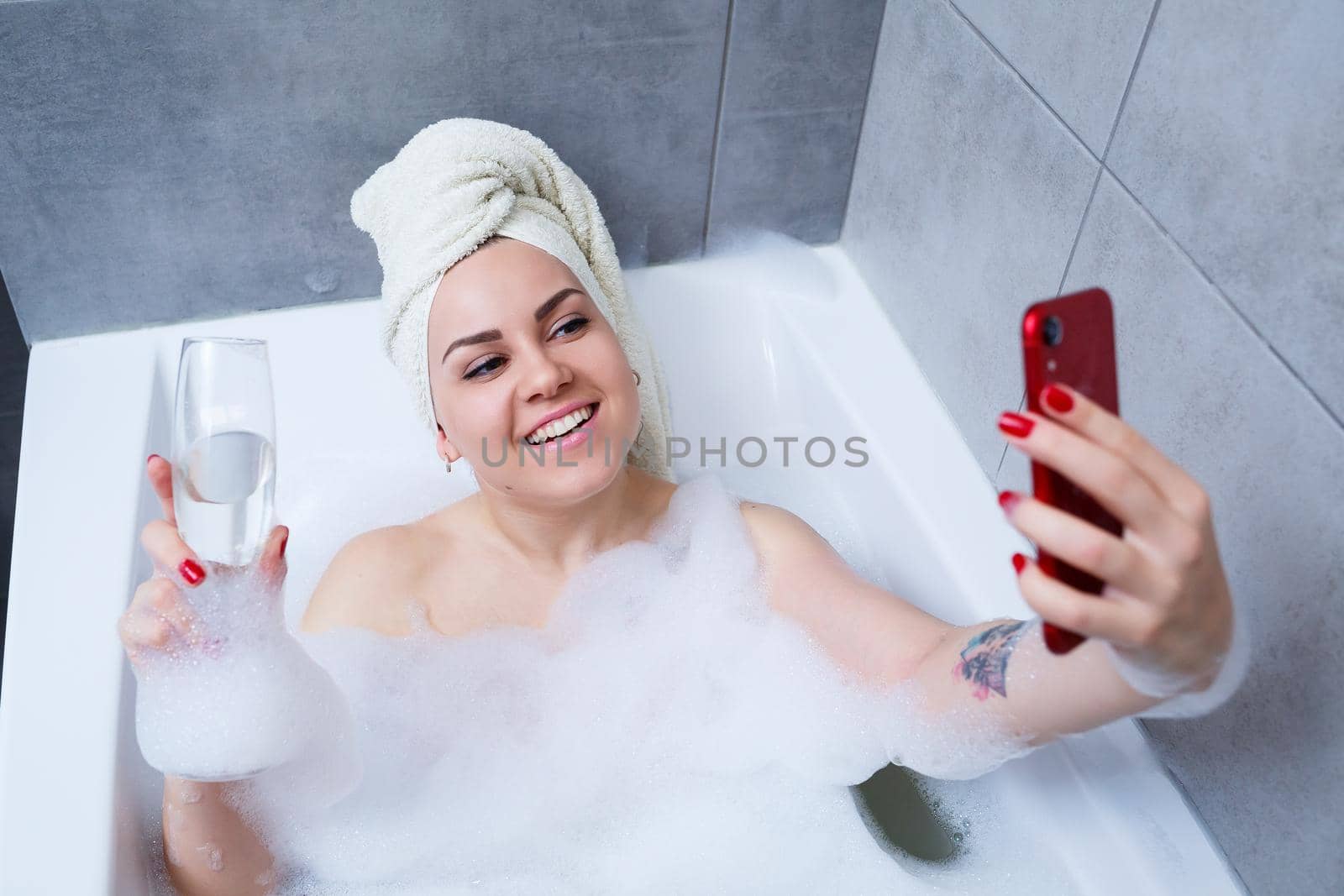 A beautiful woman with a towel on her head lies with a glass of champagne in a white bath and takes a selfie on the phone. Relax after a hard day. Relaxing spa treatment